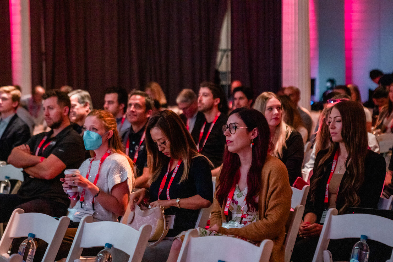 Rows of seated attendees watching the stage, some wearing masks, with pink lit columns behind them.