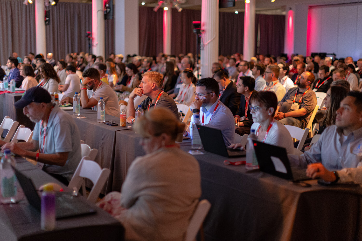 Rows of attendees in lanyards at draped tables with laptops and drinks, watching the stage.