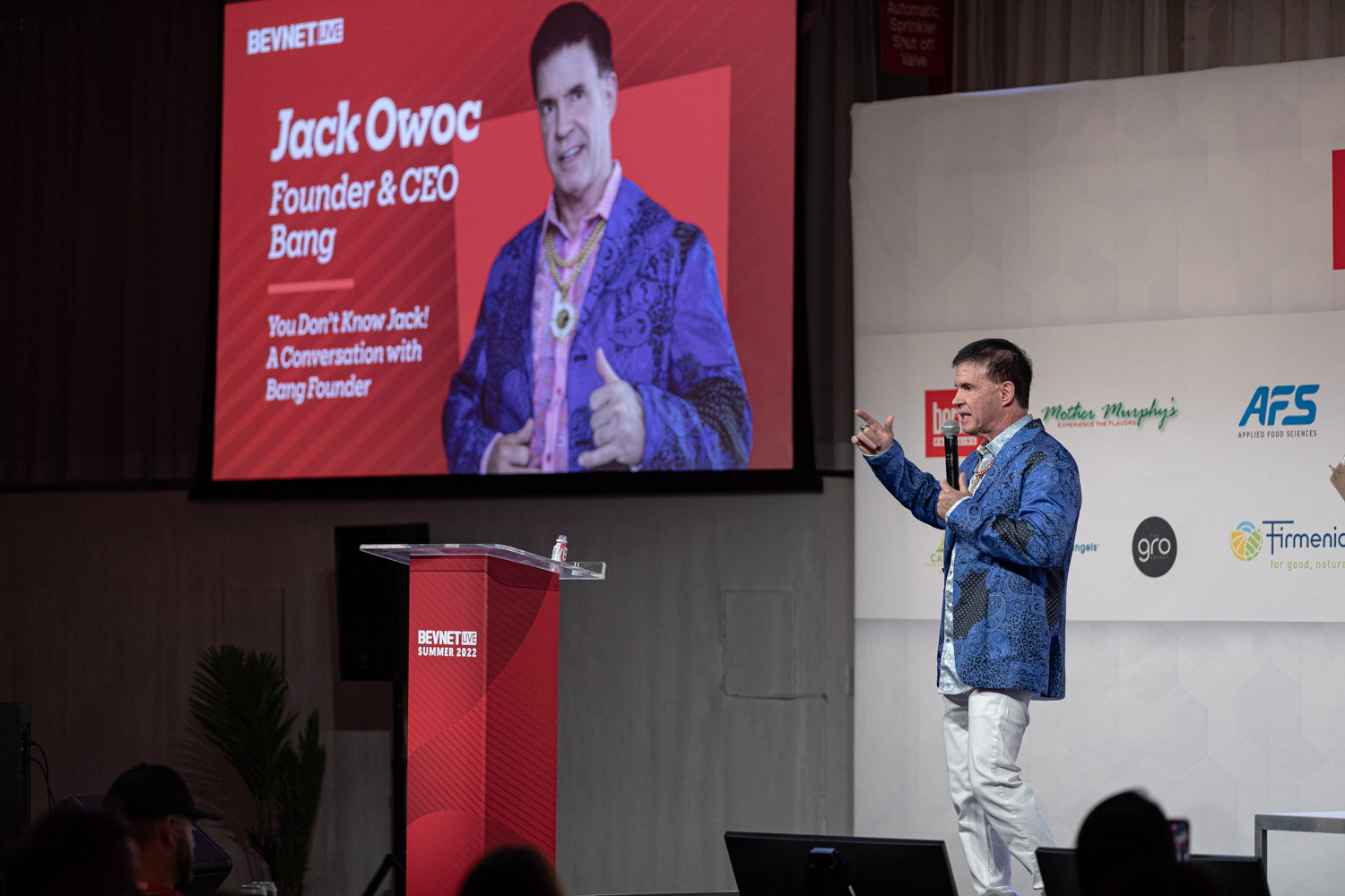 A man in a blue jacket and white pants speaks on stage next to a red podium, with a large screen reading Bang behind him.