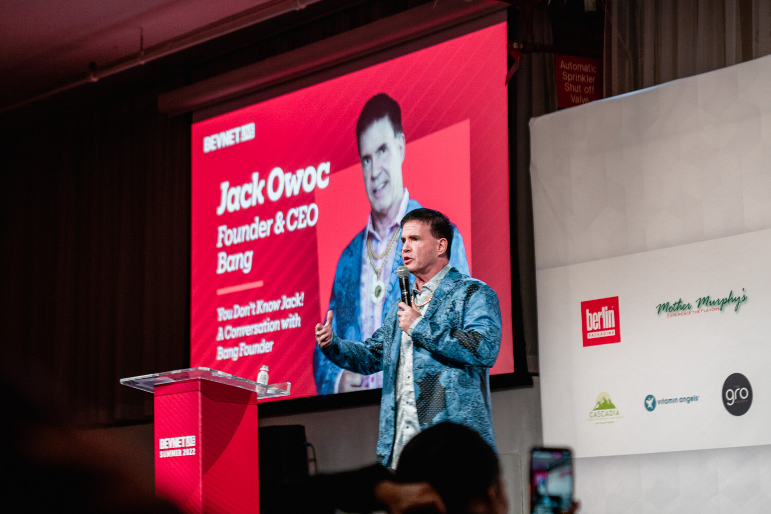 A man with a microphone stands before a red screen with the Bang session title at a lit stage.