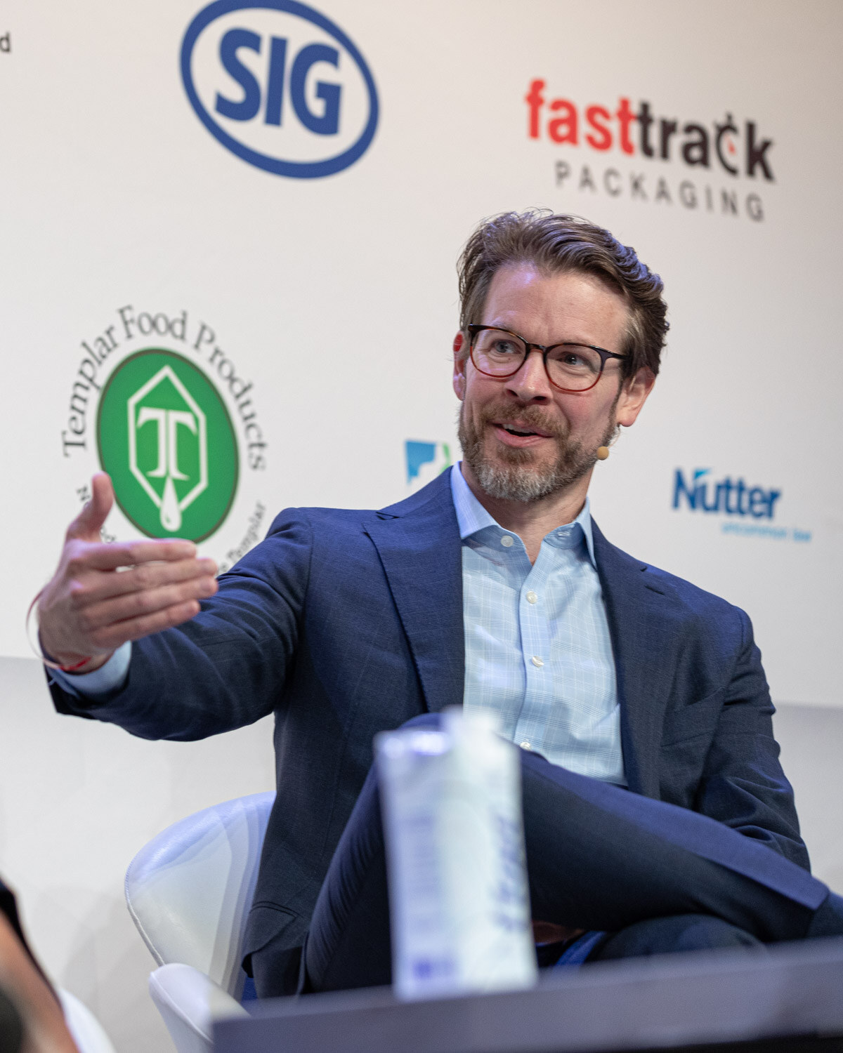 A bearded man in glasses and a navy blazer extends one hand while seated, a Flow carton on the table in front of him.
