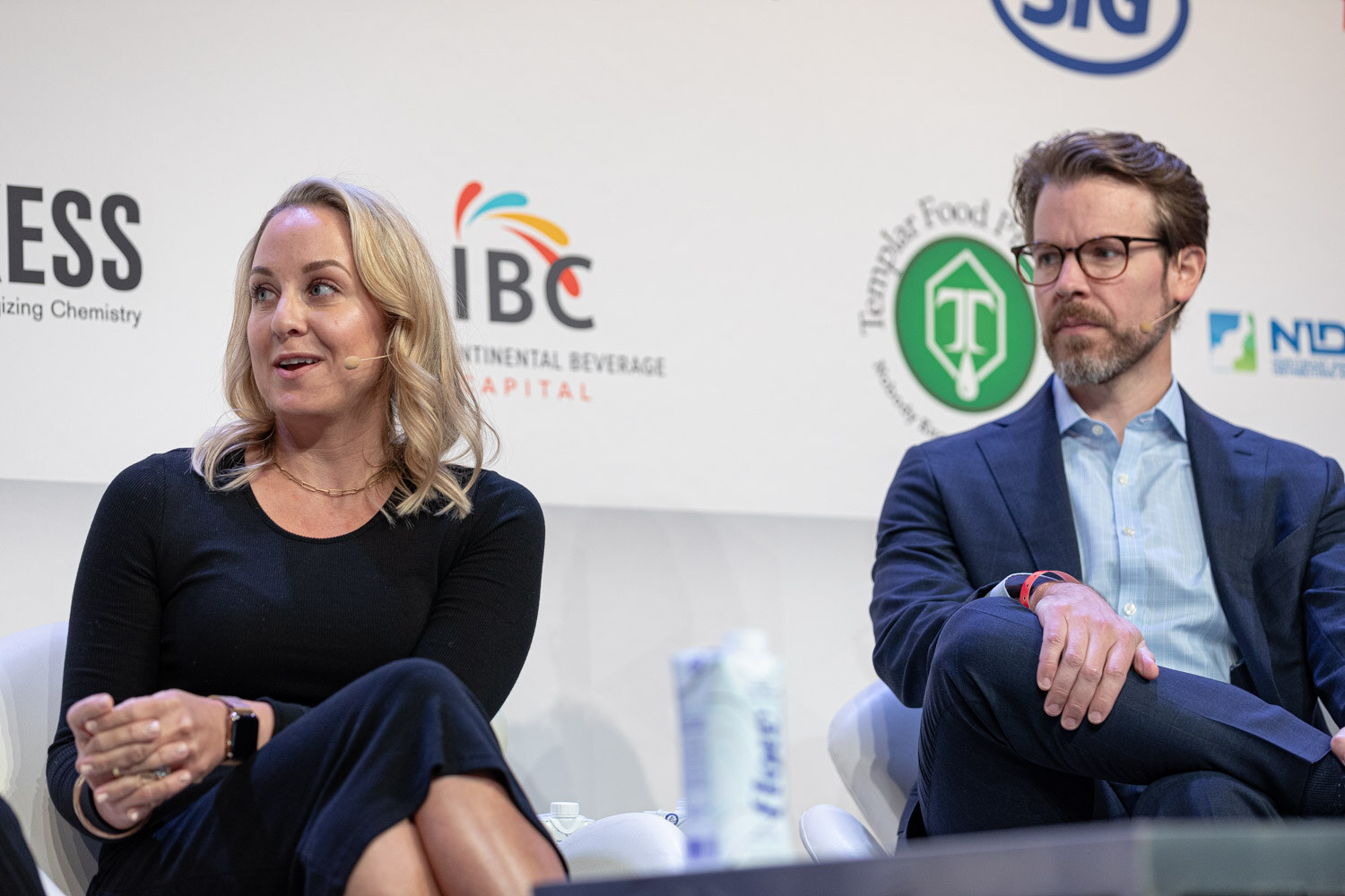 A woman with blonde hair in a black dress speaks while a bearded man in glasses and a navy blazer listens.