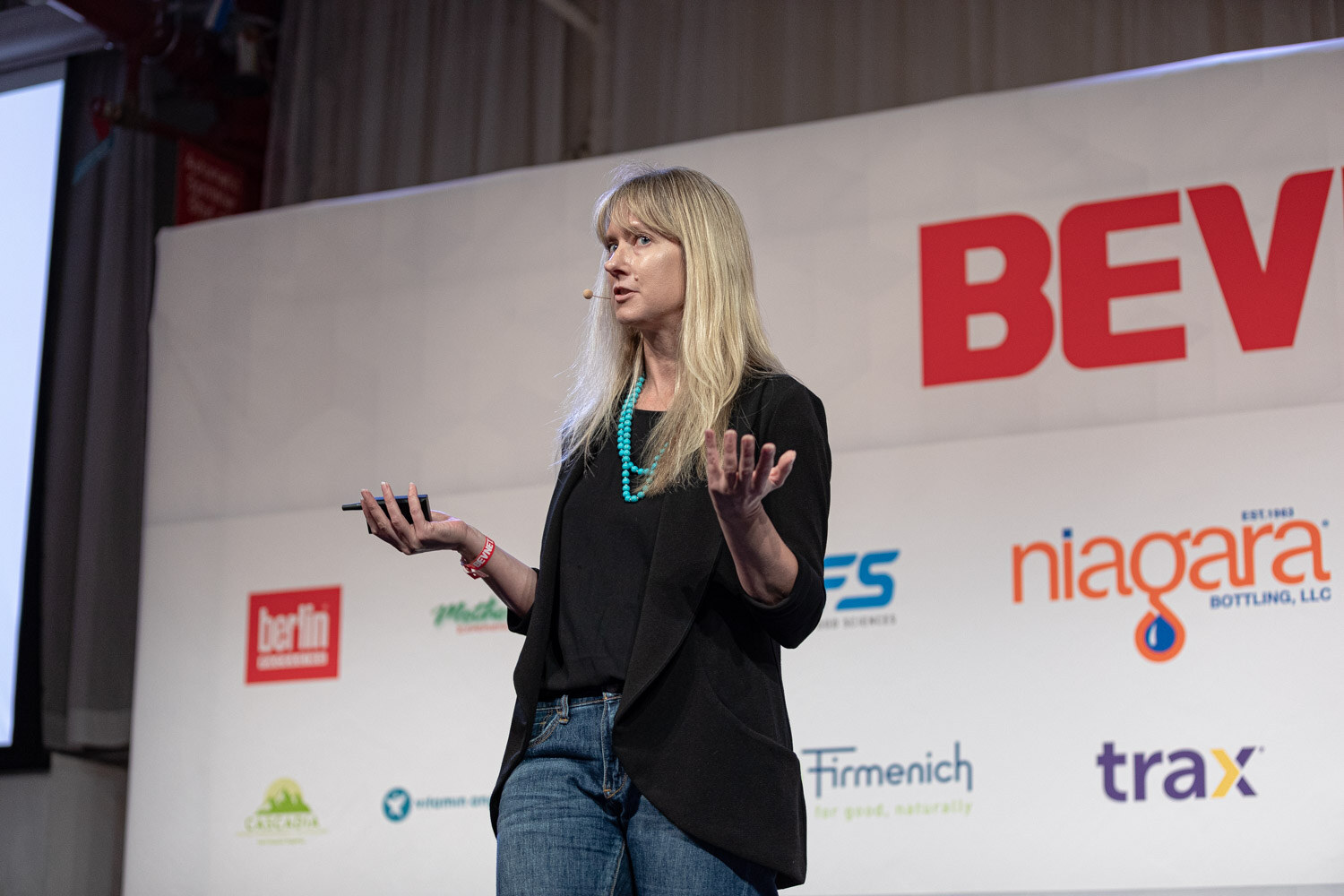 A woman with long blonde hair gestures with a phone in hand in front of a BevNET Live backdrop.