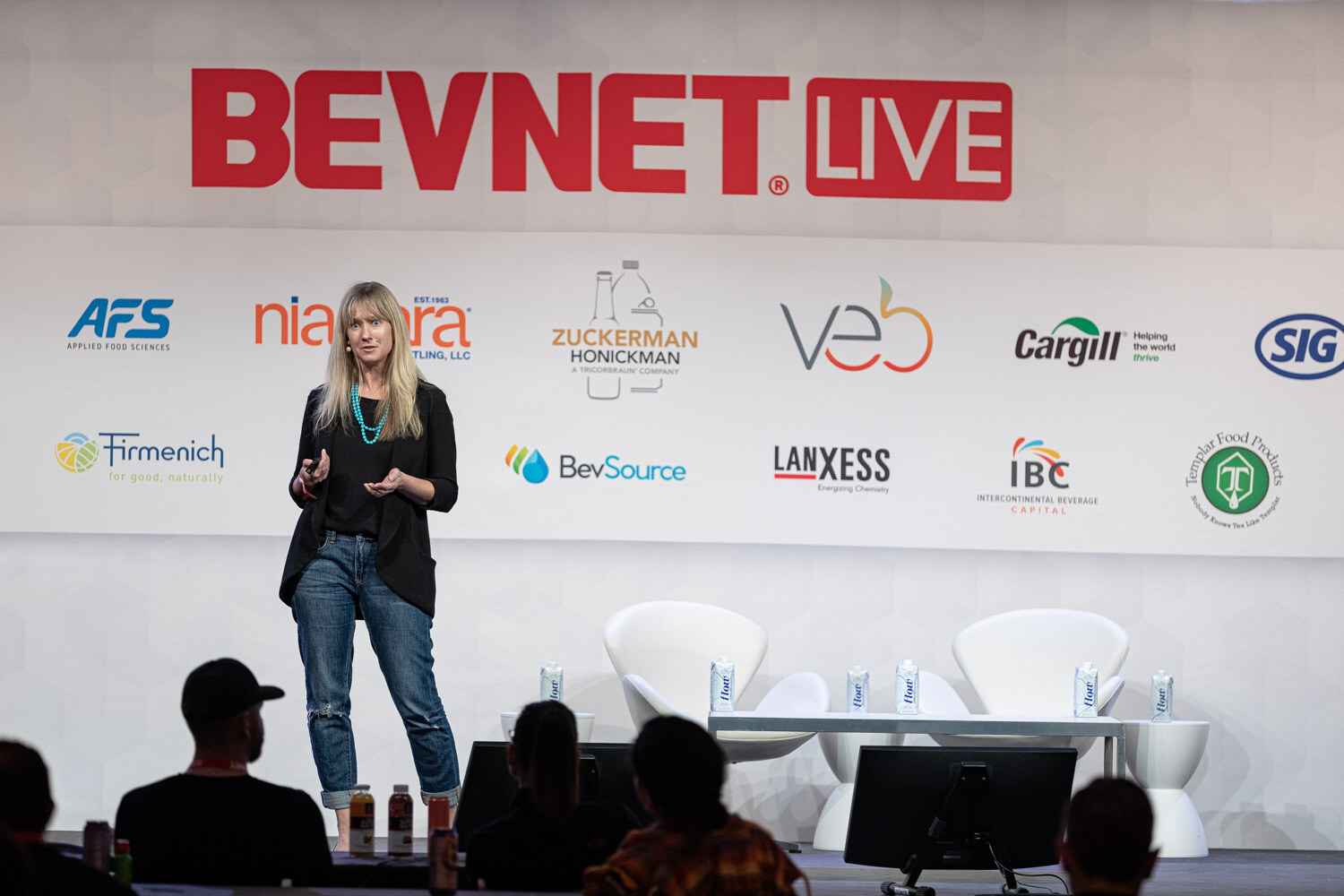 A woman with long blonde hair and a black blazer stands on a stage in front of a BevNET Live backdrop with sponsor logos, with audience heads in the foreground.