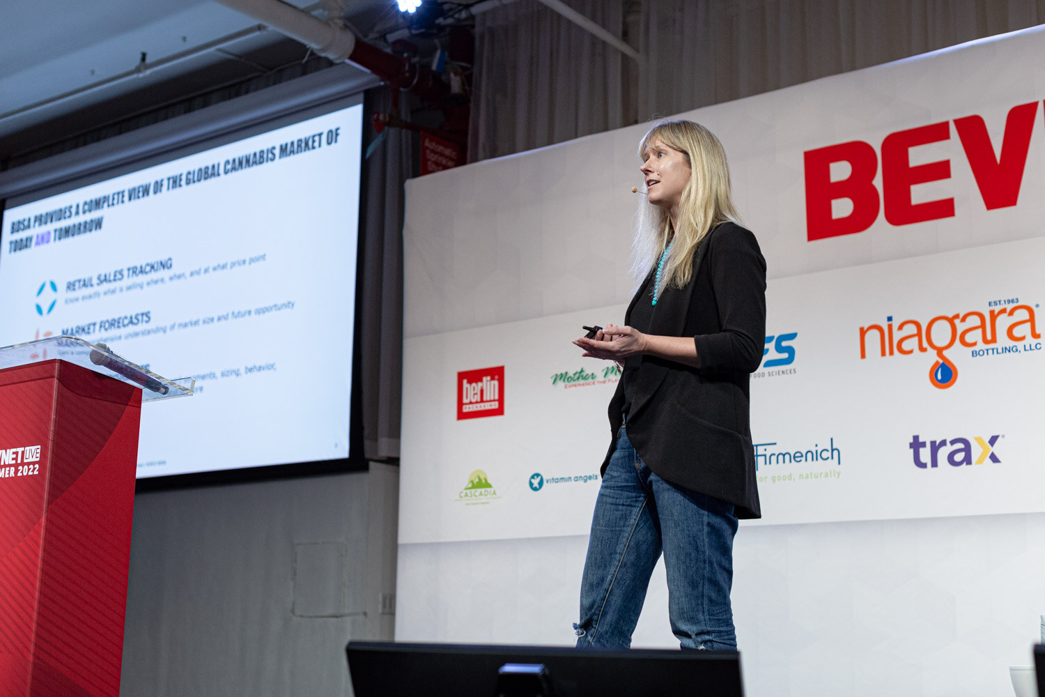 A woman in a black blazer stands next to a podium and a projector screen with text about retail sales tracking and market forecasts.
