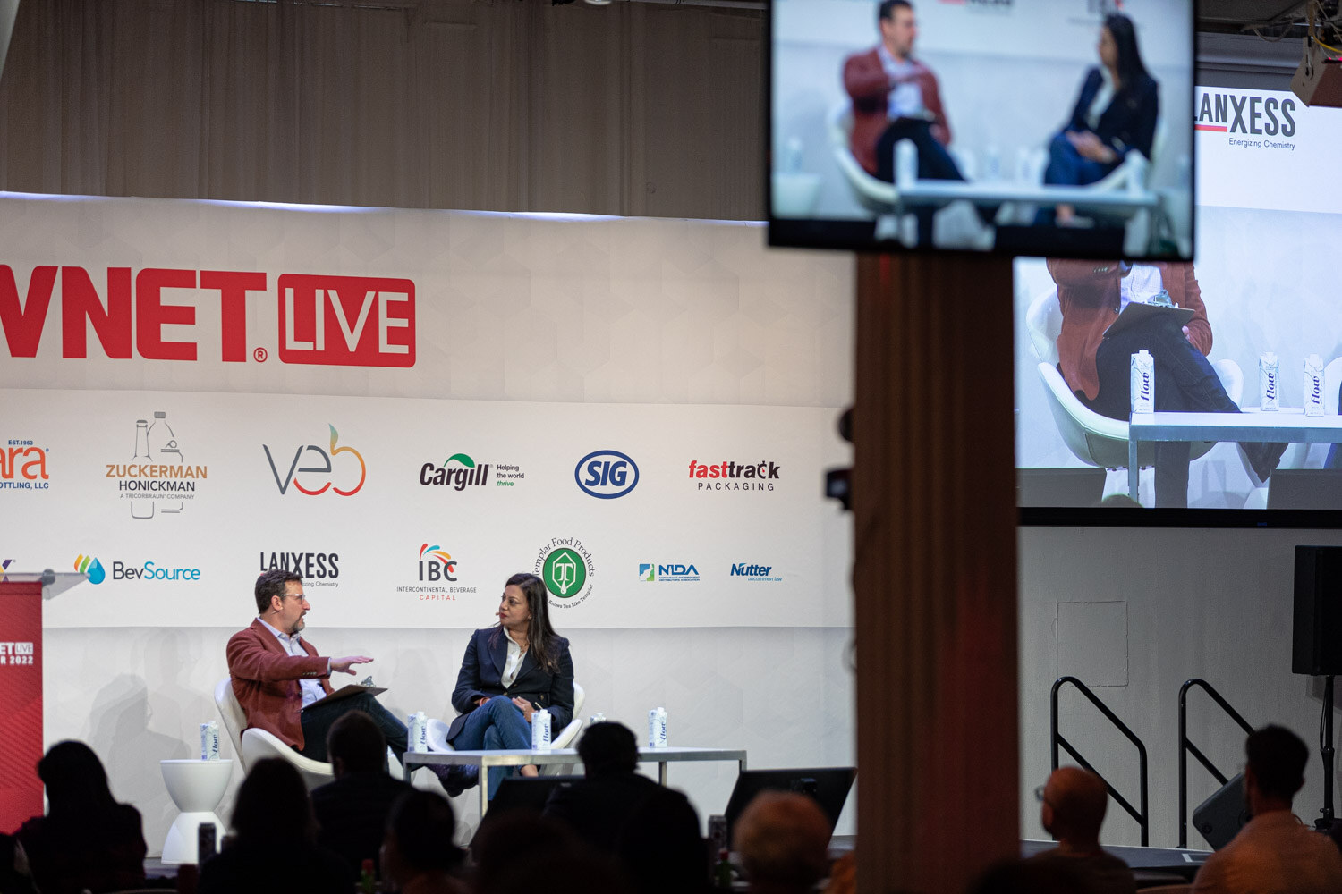 Wide view of a man in a rust jacket and a woman in a navy blazer seated on a stage below a BevNET Live sign, a large screen beside them.
