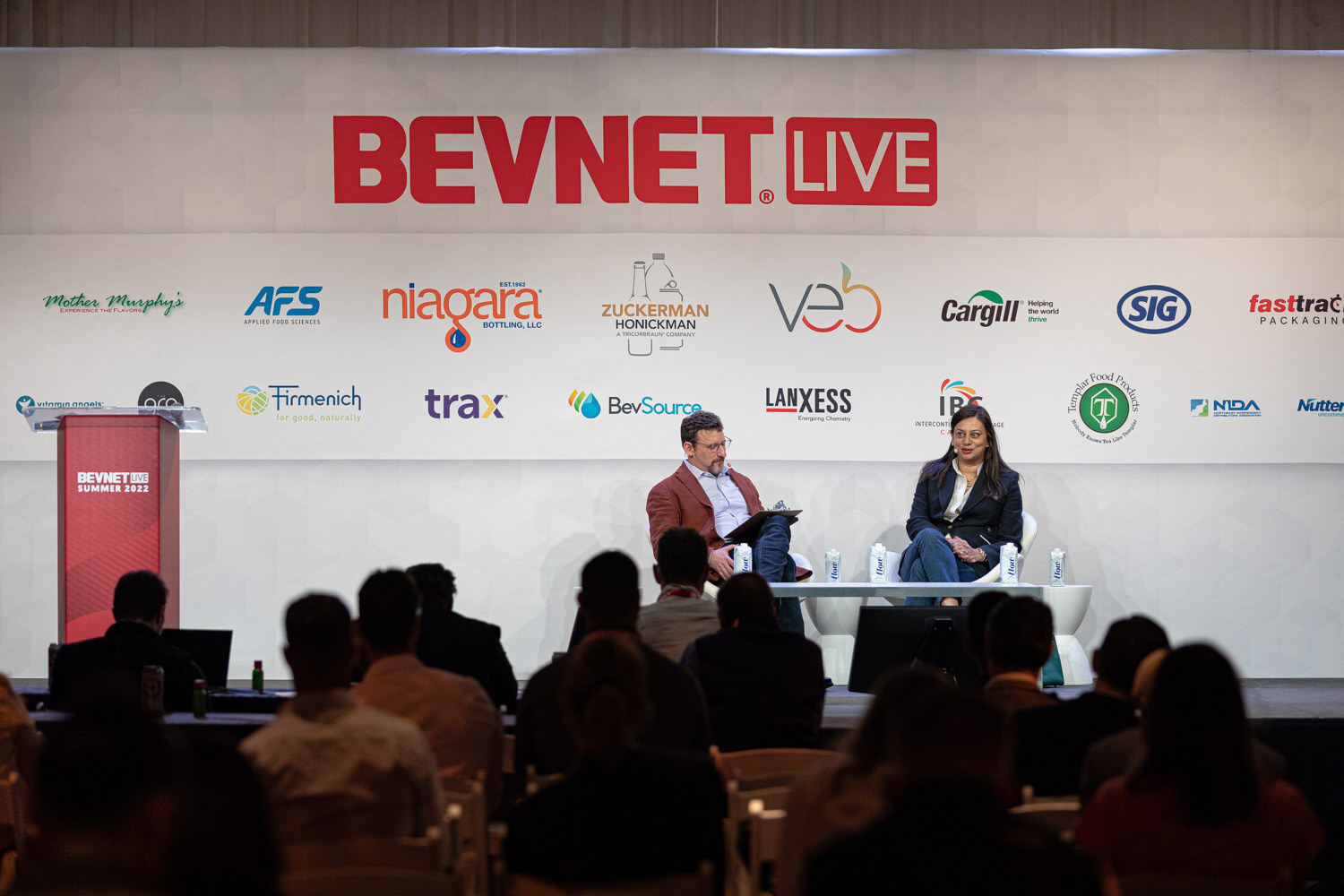 A man in a rust jacket and a woman in a navy blazer sit in white chairs on a stage below a BevNET Live backdrop.