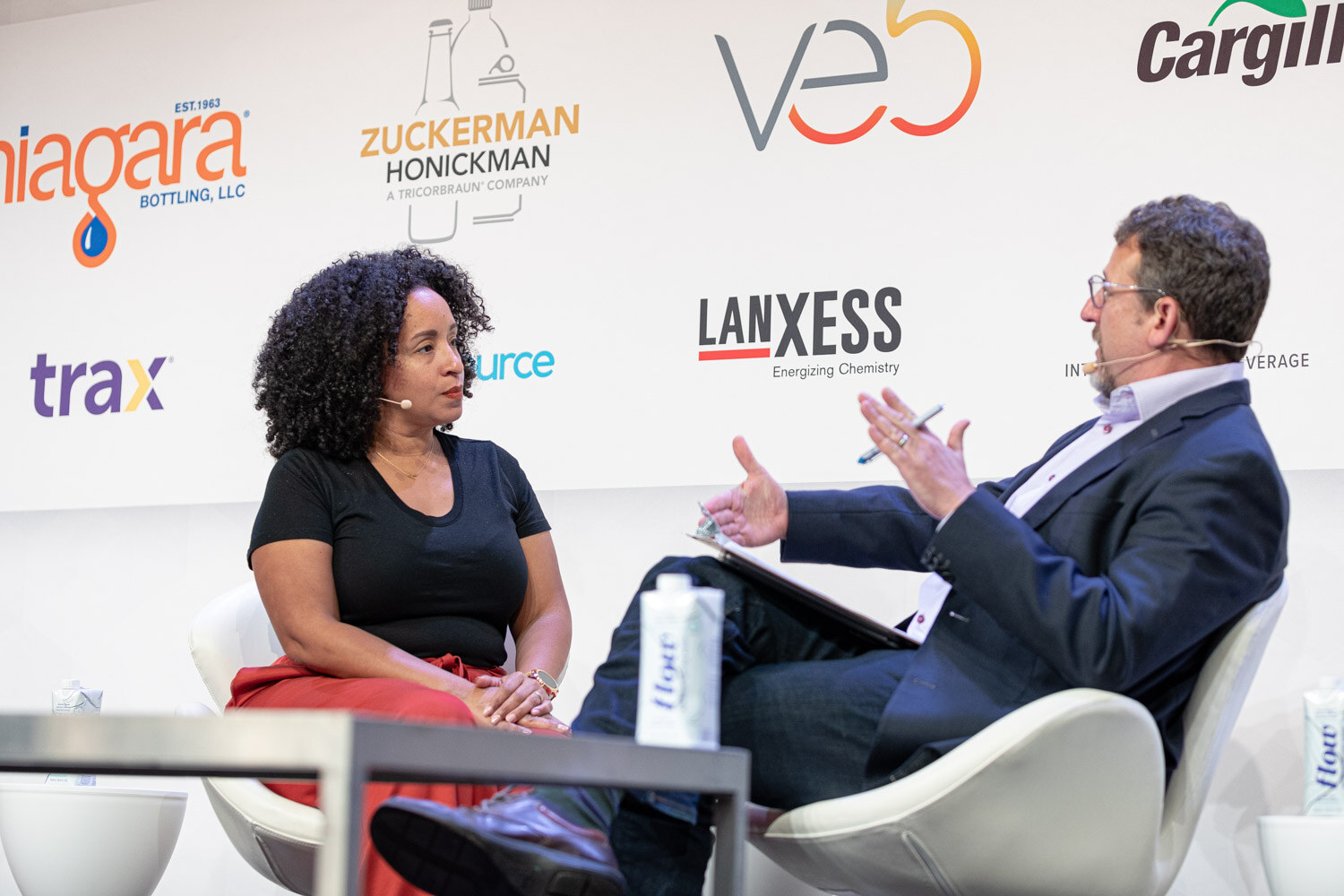 A woman in a black shirt and a man in a navy blazer with a notepad talk in white chairs in front of a sponsor-covered backdrop.