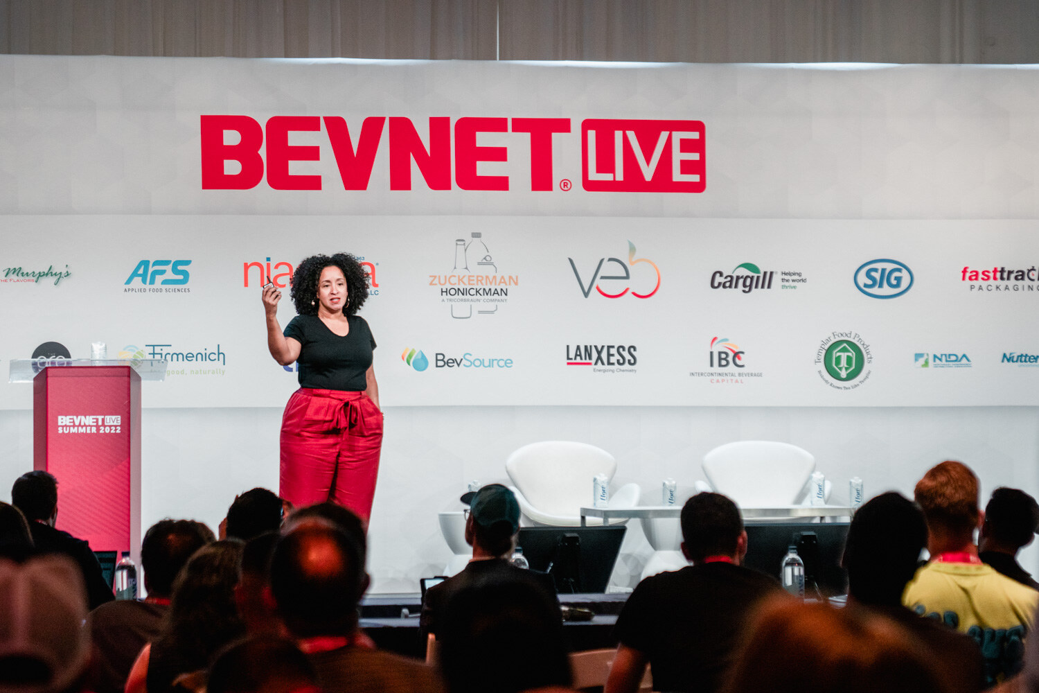A woman in a black shirt and red pants stands on the stage with attendees in lanyards seen from behind.