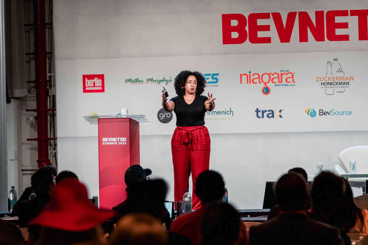 A woman in a black shirt and red pants speaks on stage next to a red podium reading BevNET Live Summer 2022.