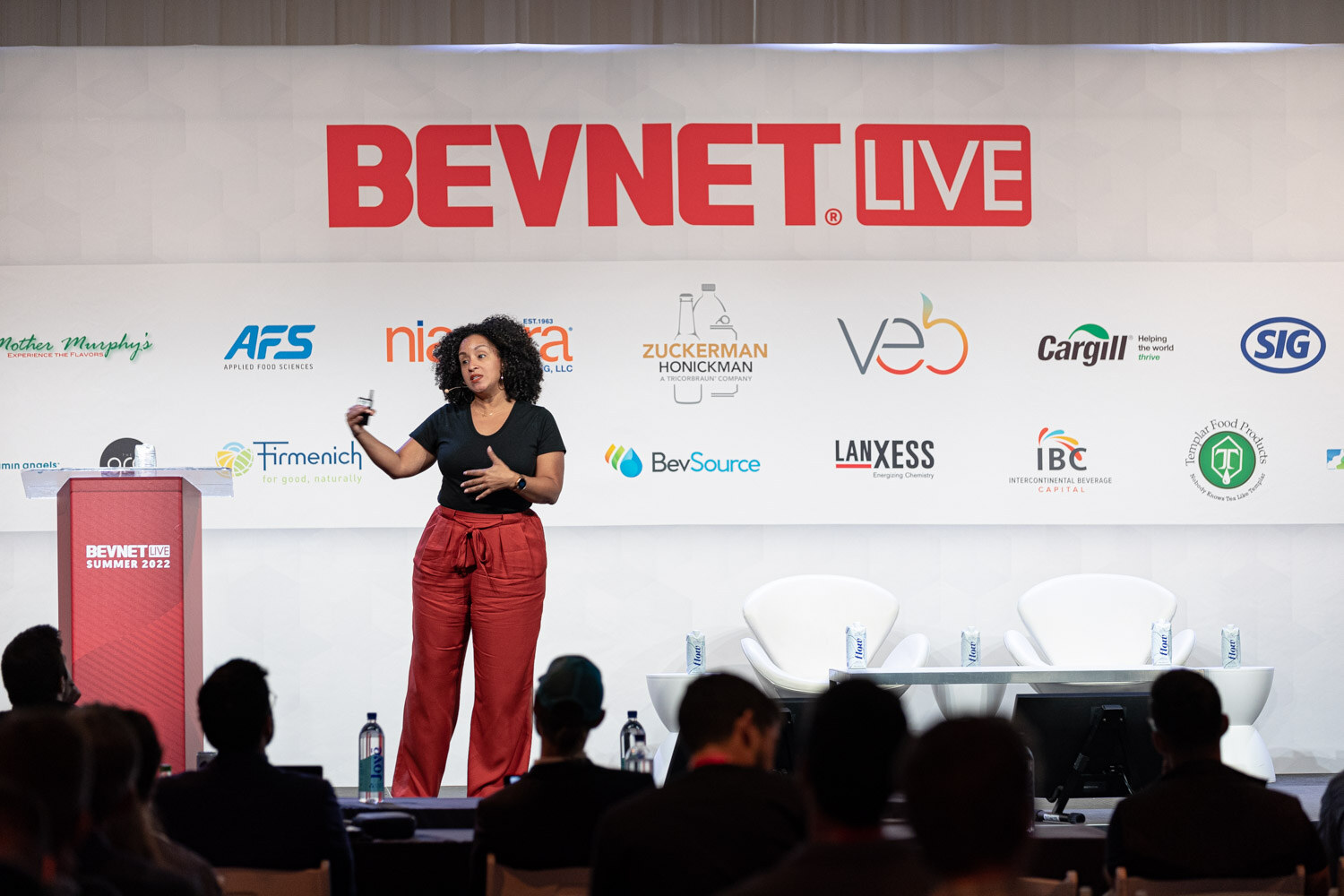A woman in a black shirt and red pants holds a clicker on a stage below the BevNET Live sign, audience silhouettes in the foreground.