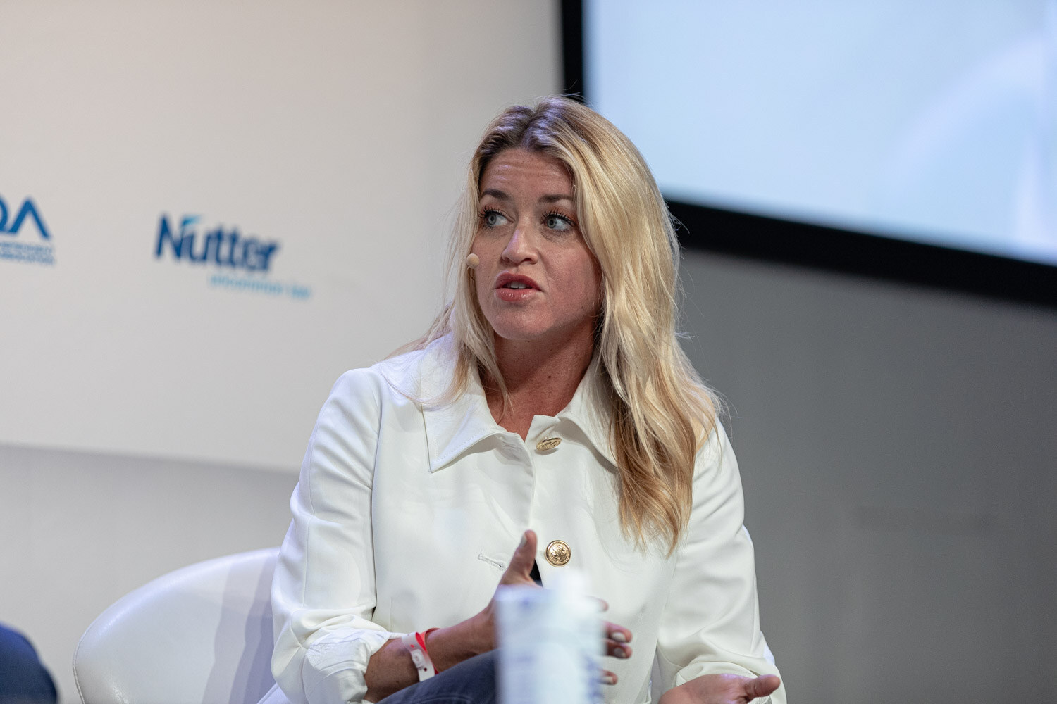 A woman with blonde hair in a white jacket sits with a headset microphone in front of sponsor logos.