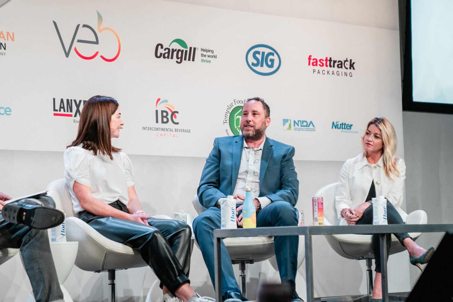 A woman in a white blouse smiles at a man in a teal blazer who talks while a woman in a white jacket listens, in front of sponsor logos.