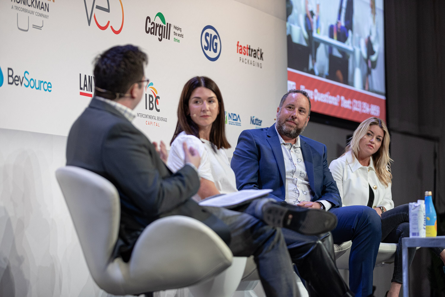 Four people seated in white chairs on stage, a moderator with his back to the camera and three panelists facing him.