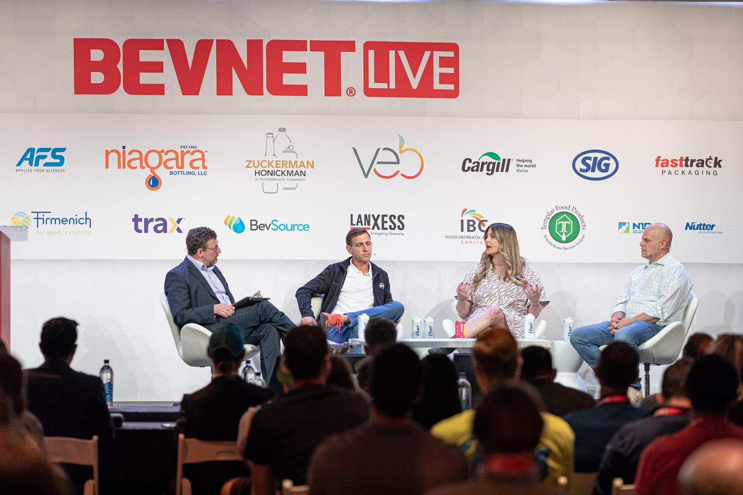Four people seated on a stage in white chairs under a BevNET Live sign with audience members' heads in the foreground.