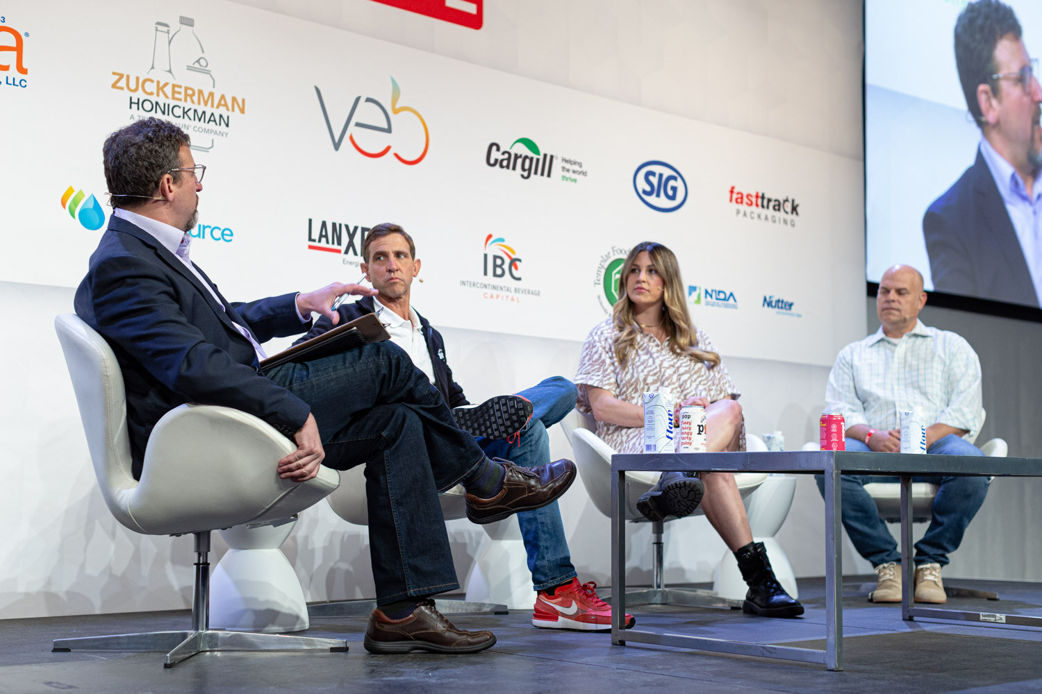 Four people seated on white swivel chairs on stage, a moderator in the foreground facing a man in a quarter-zip, a woman in a patterned dress and a man in a checked shirt.