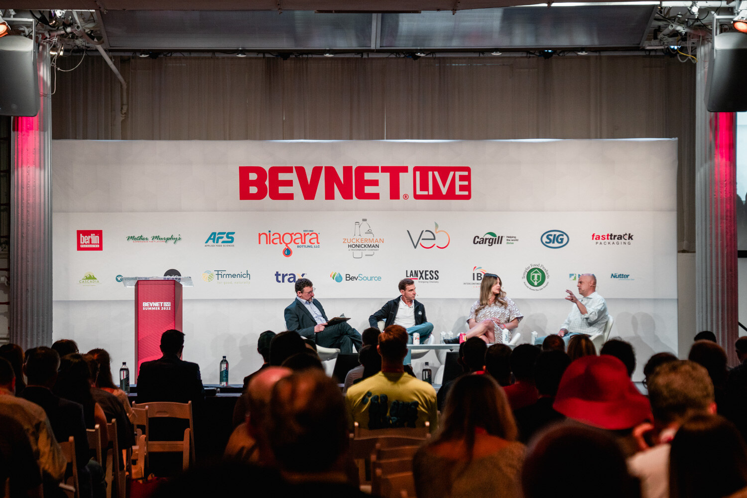 Wide view of the stage with four seated speakers and an audience of attendees in the foreground.