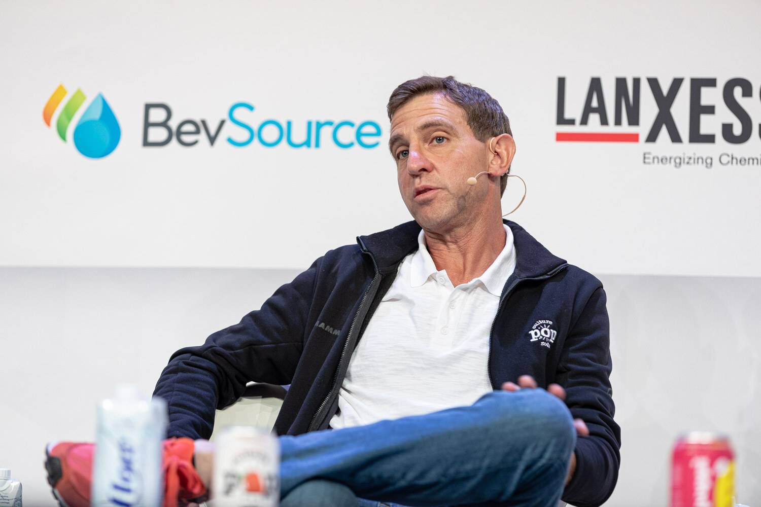 A man in a navy pullover and white shirt sits with legs crossed, soda cans in the foreground and a BevSource logo behind.