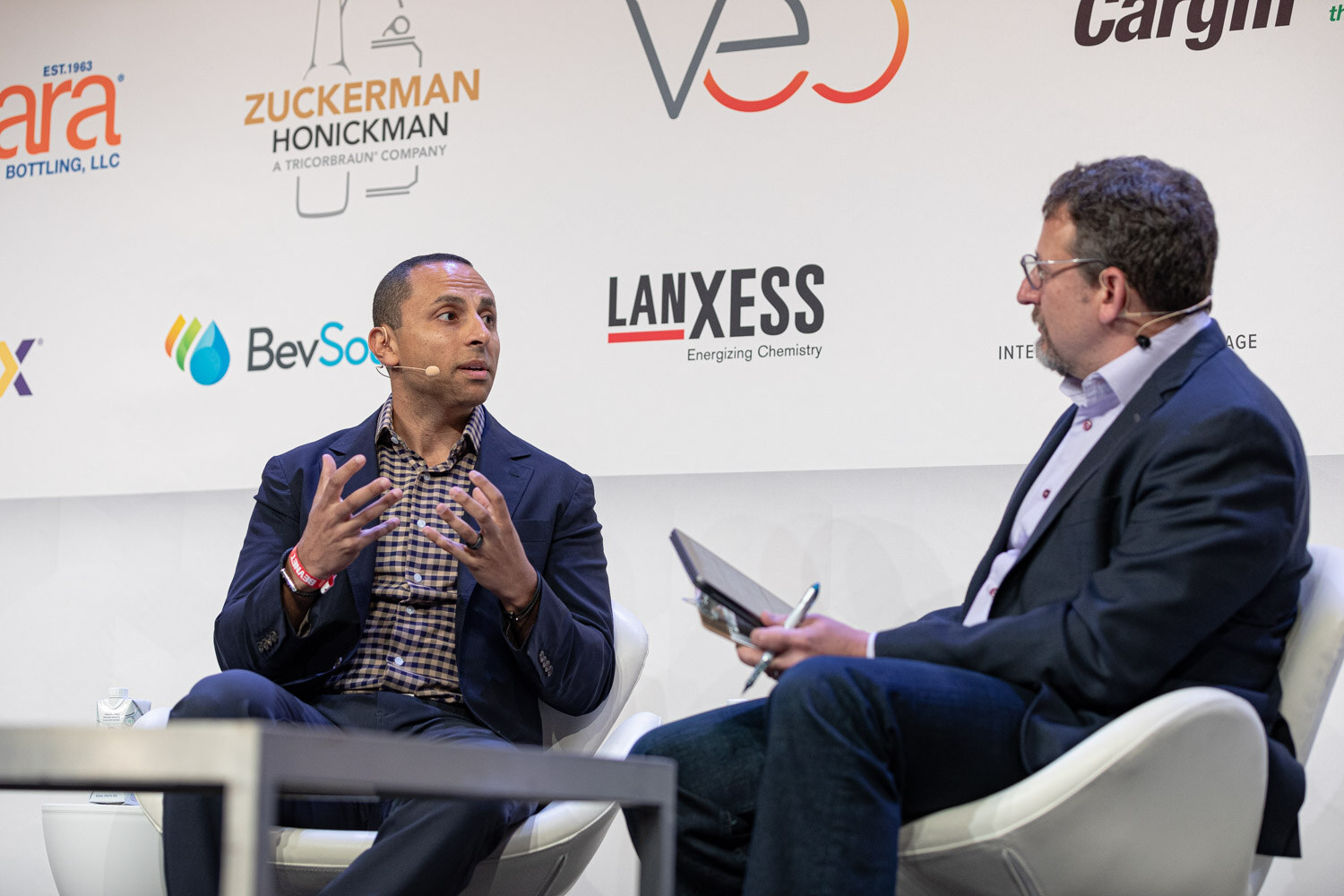 A man in a navy blazer and a man holding a tablet and pen sit in white chairs, talking, with sponsor logos behind them.