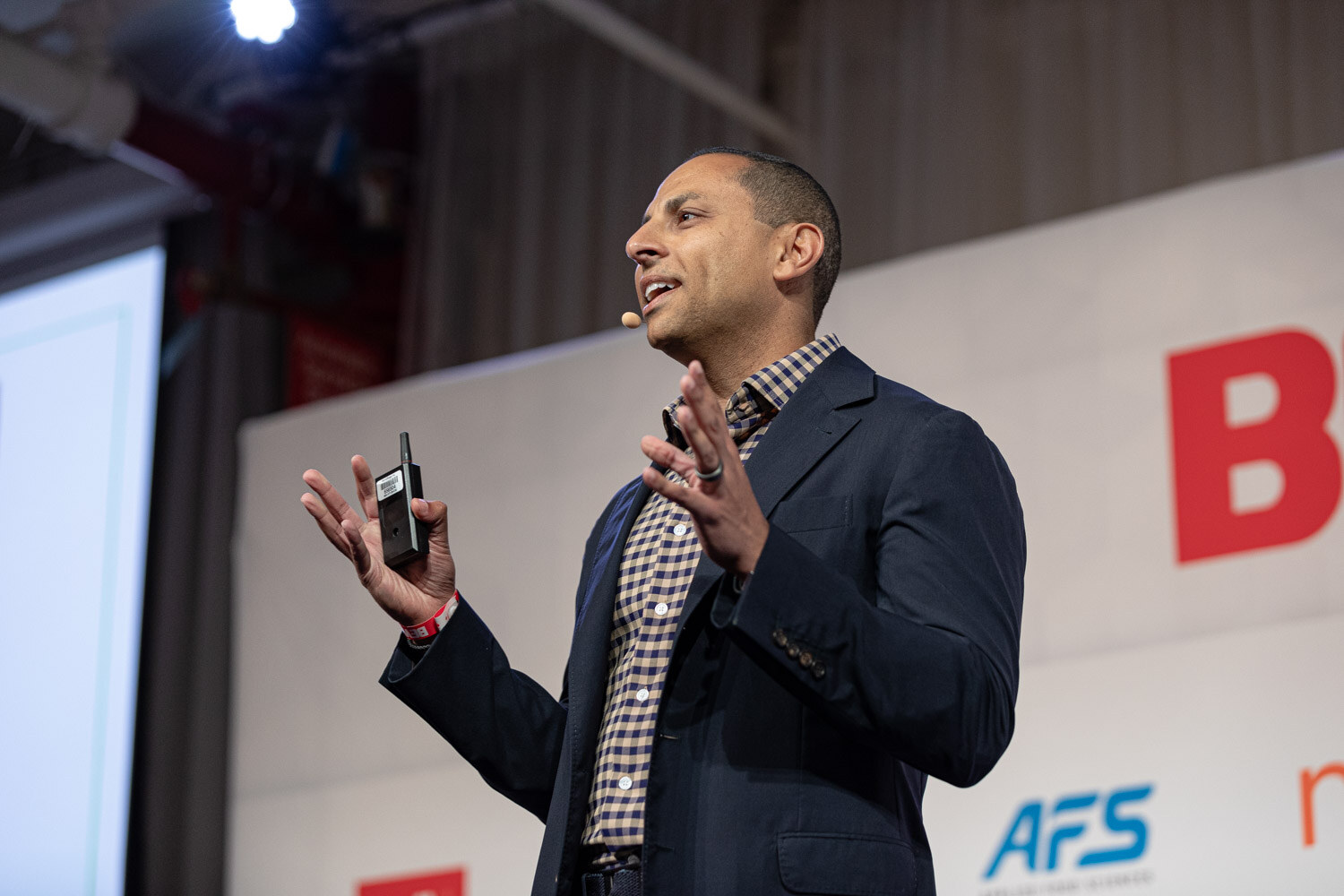 A man in a navy blazer and checked shirt gestures with raised hands on stage, with AFS signage behind him.