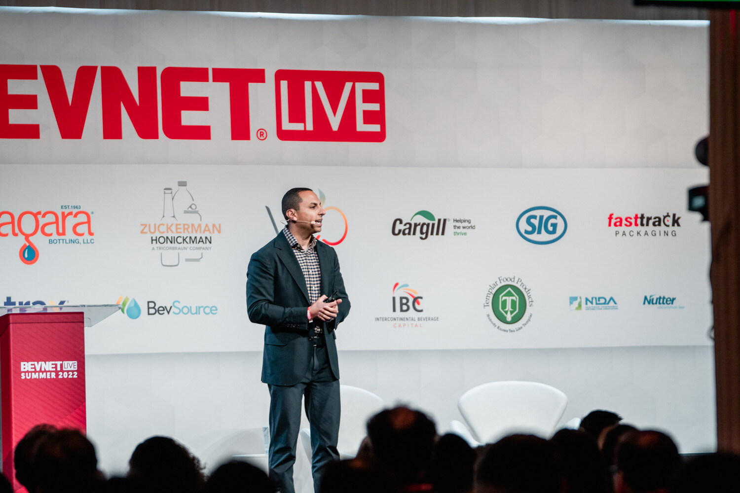 A man in a navy blazer and checked shirt stands in front of a BevNET Live backdrop beside a red podium.