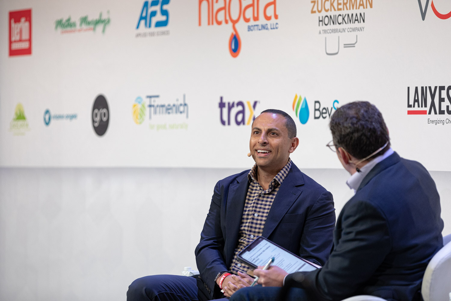 A man in a navy blazer and checked shirt smiles while a moderator with a tablet sits in the foreground.