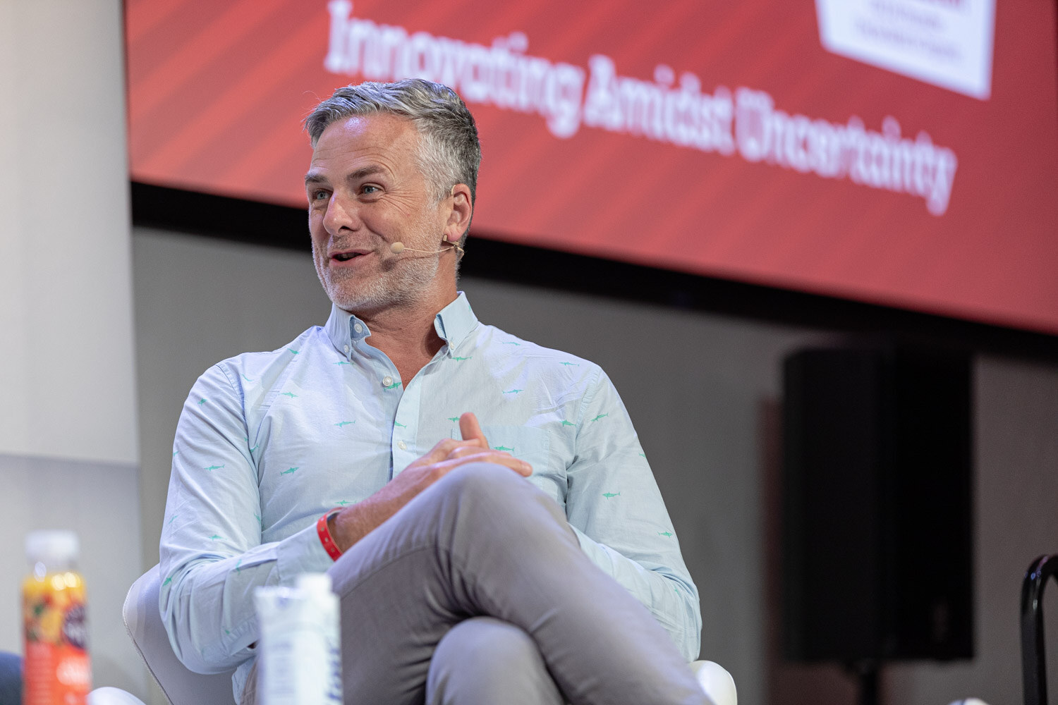 A man with gray hair in a light blue shirt sits with his legs crossed in front of a red screen reading Innovating Amidst Uncertainty.