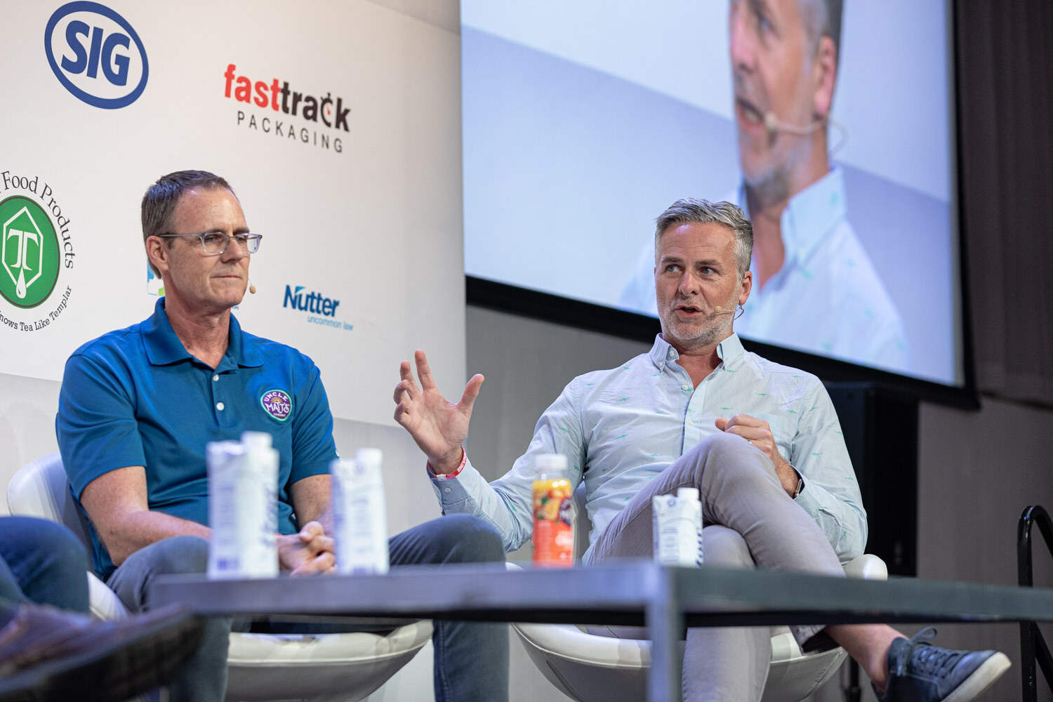 A man in a blue polo and a man in a light blue shirt sit in white chairs with cartons and a juice bottle on the table.