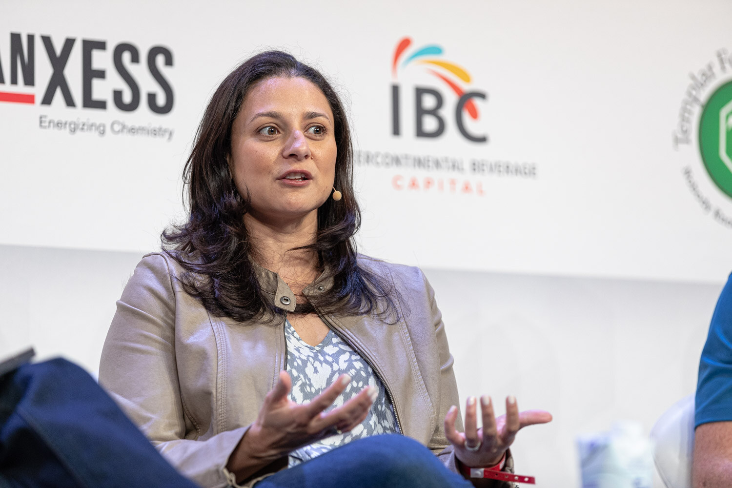 A woman with dark hair in a tan jacket gestures with both hands in front of IBC and Lanxess signs.