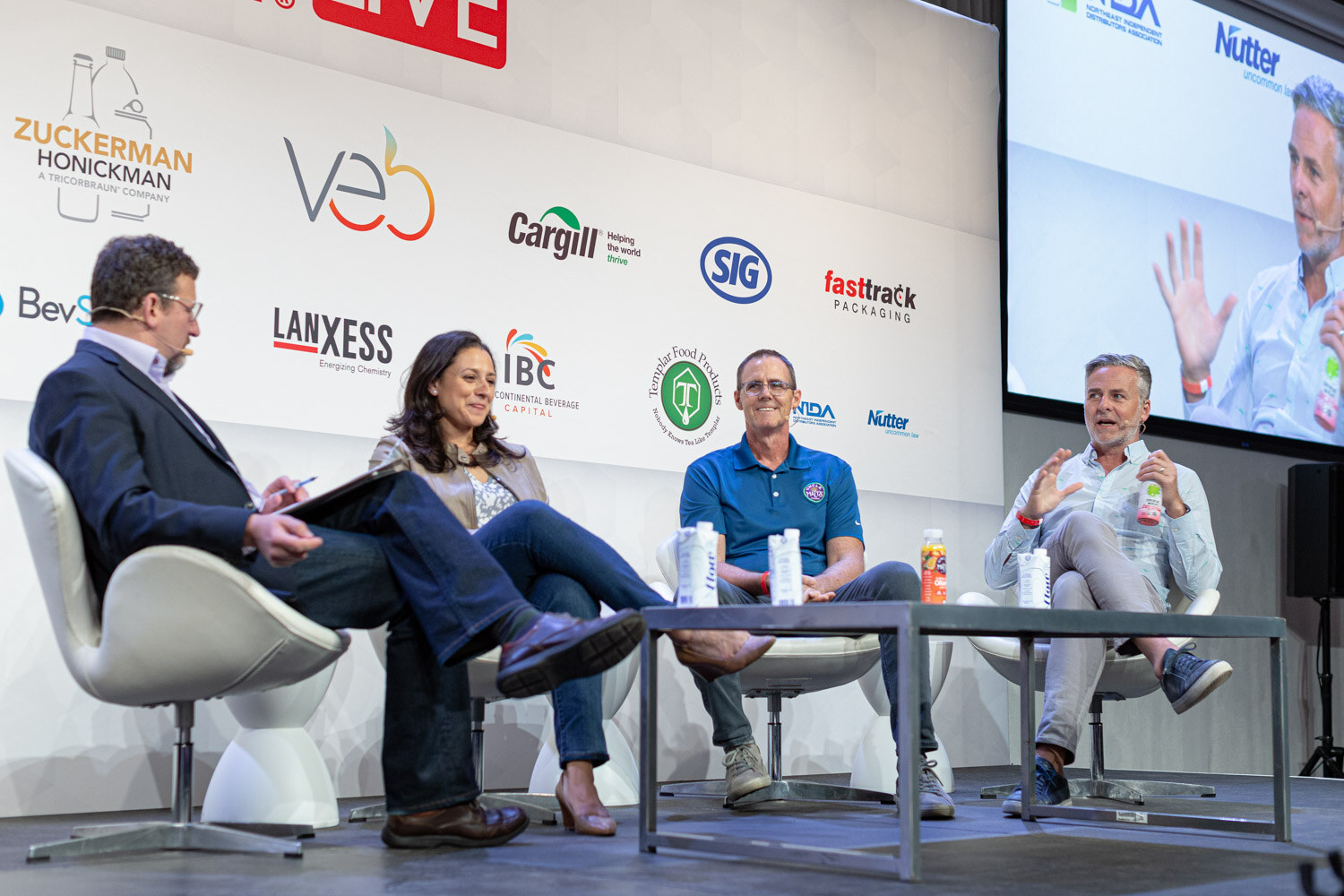 Four people sit in a low semicircle on stage, a man holding a tablet facing the three panelists, with a screen showing a close-up behind them.