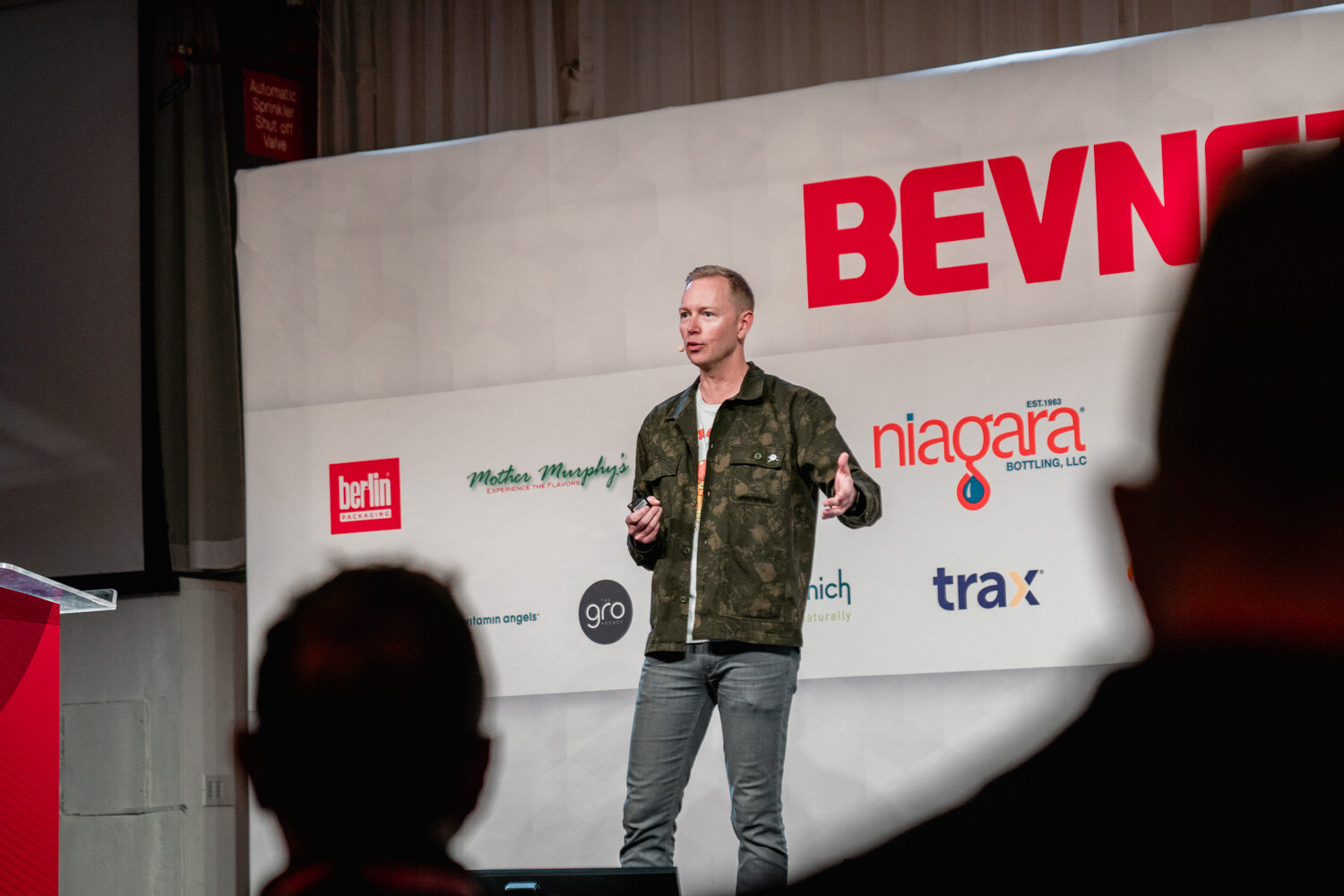 A man in a camouflage jacket and gray jeans stands holding a clicker and gesturing in front of a BevNET Live backdrop.