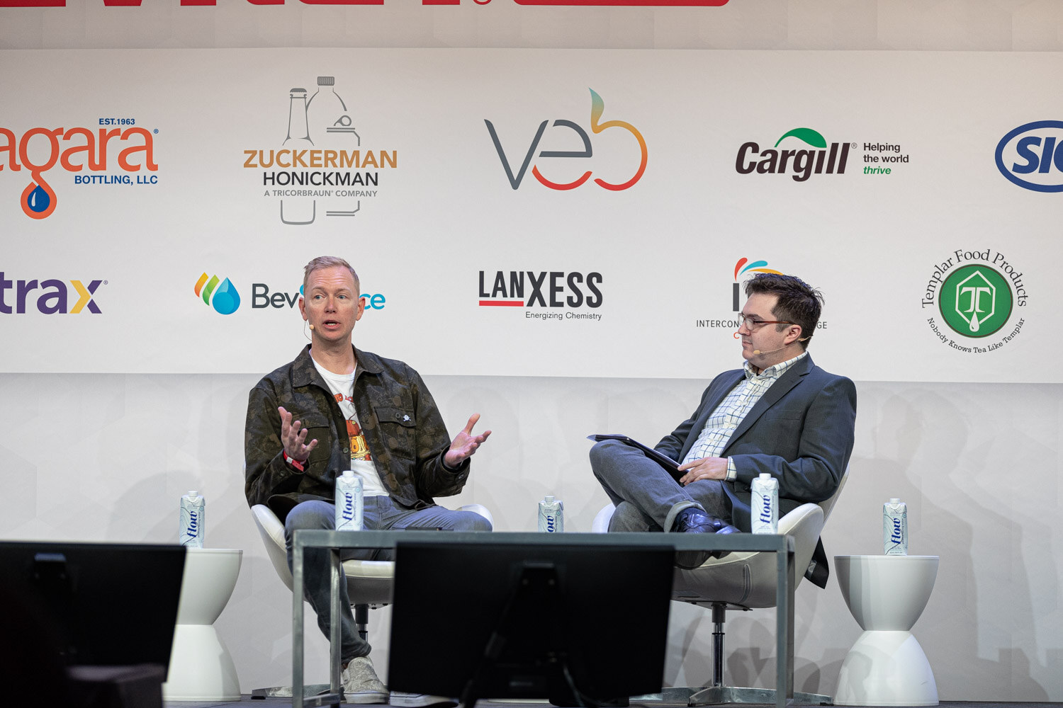 A man in a camouflage jacket with open hands talks with a man in a gray blazer holding a tablet, in front of sponsor logos.