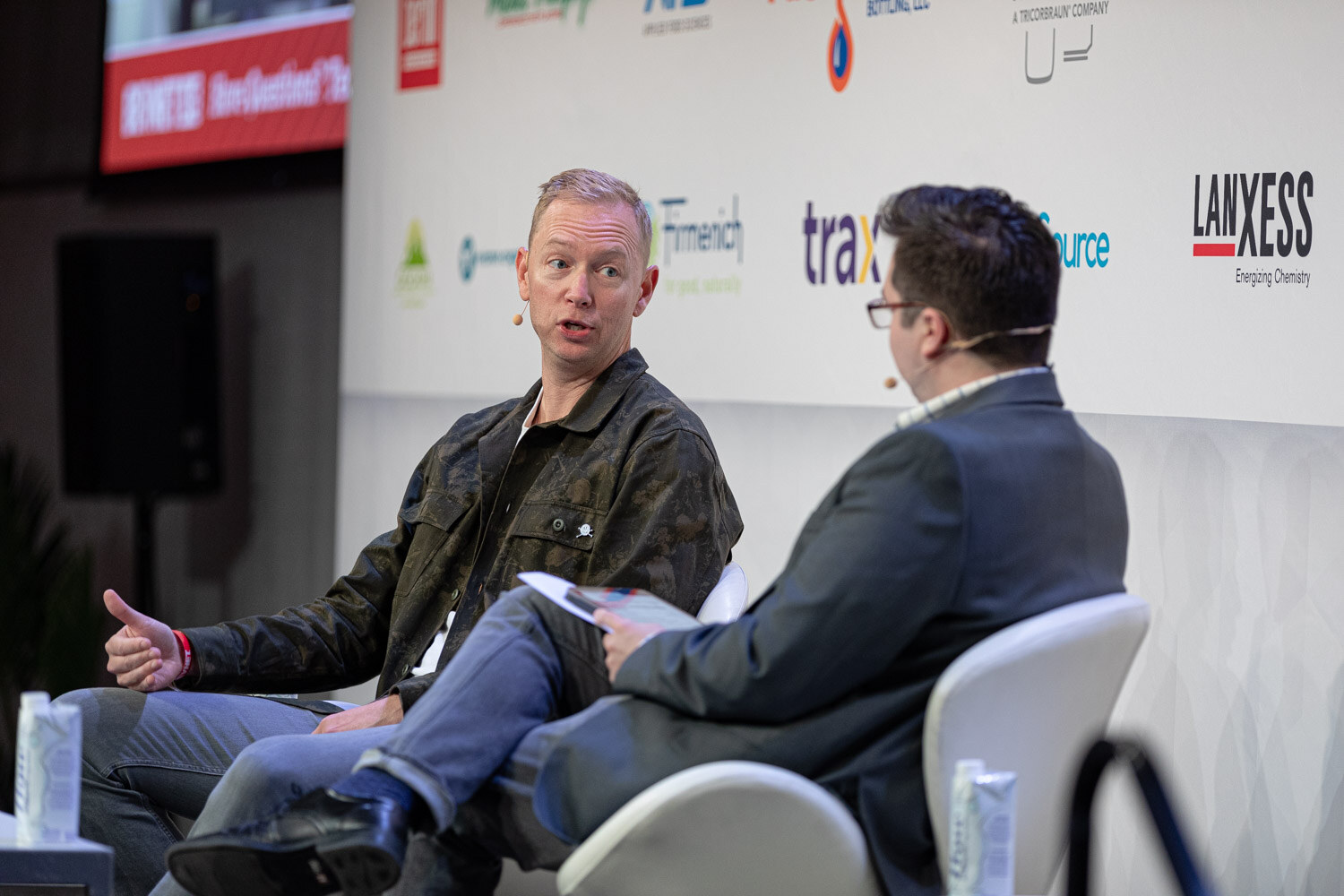 A man in a camouflage jacket and a man in a navy blazer with a tablet sit in white chairs in front of sponsor logos.