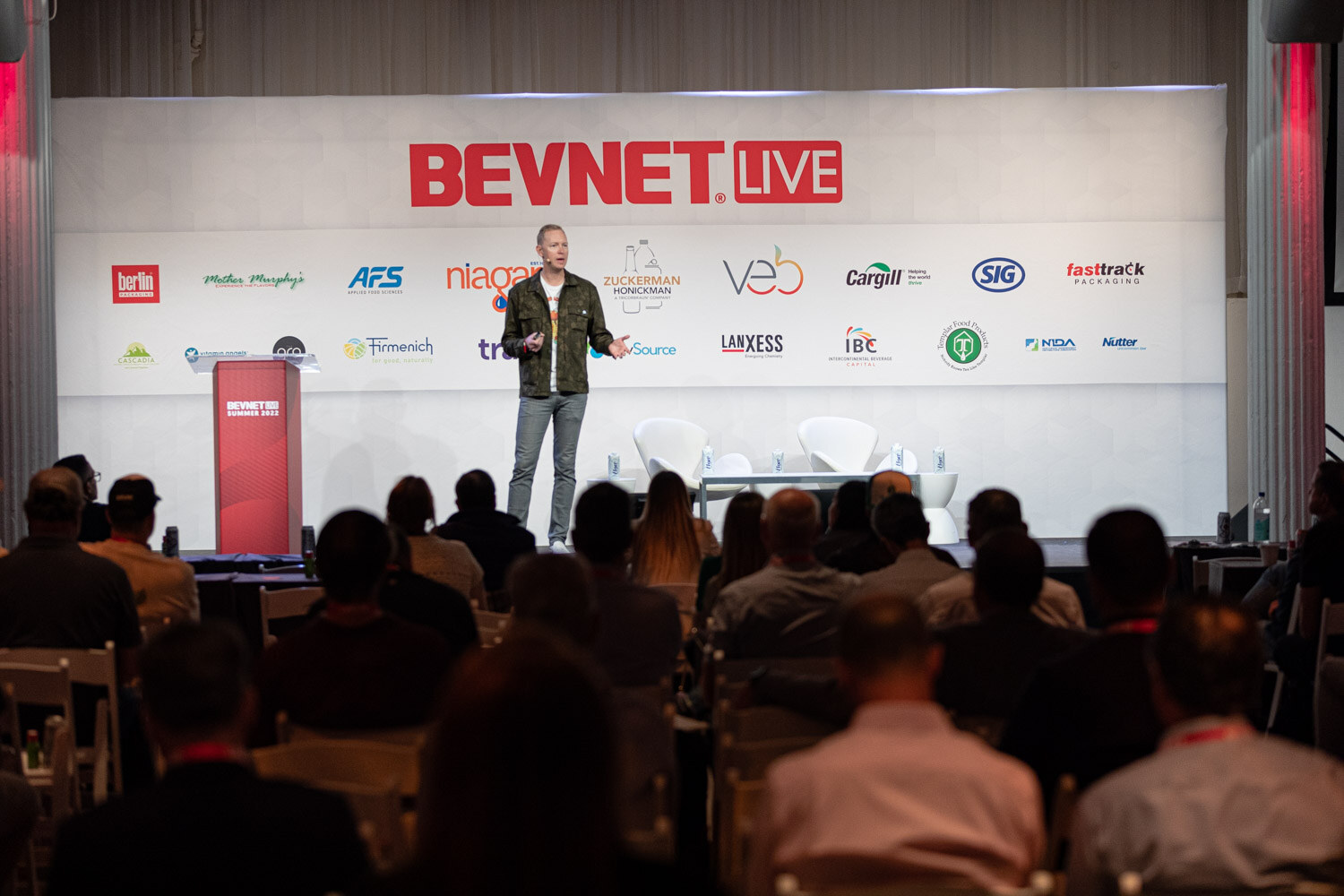 Wide view of a man in a camouflage jacket on the BevNET Live stage with silhouettes of attendees in front.