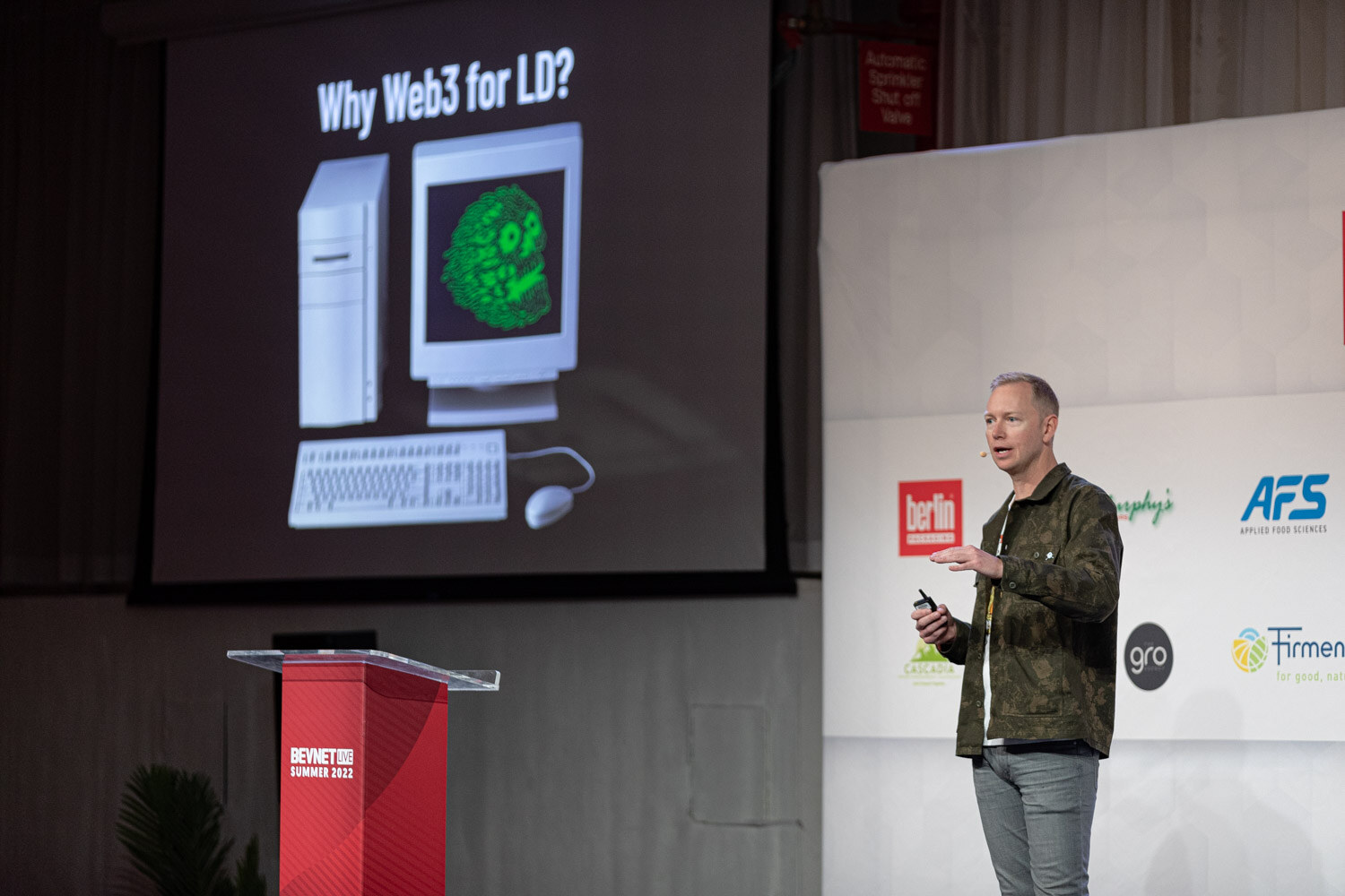 A man in a camouflage jacket stands on stage next to a red podium with a projector screen showing an old computer and a green skull on its monitor.