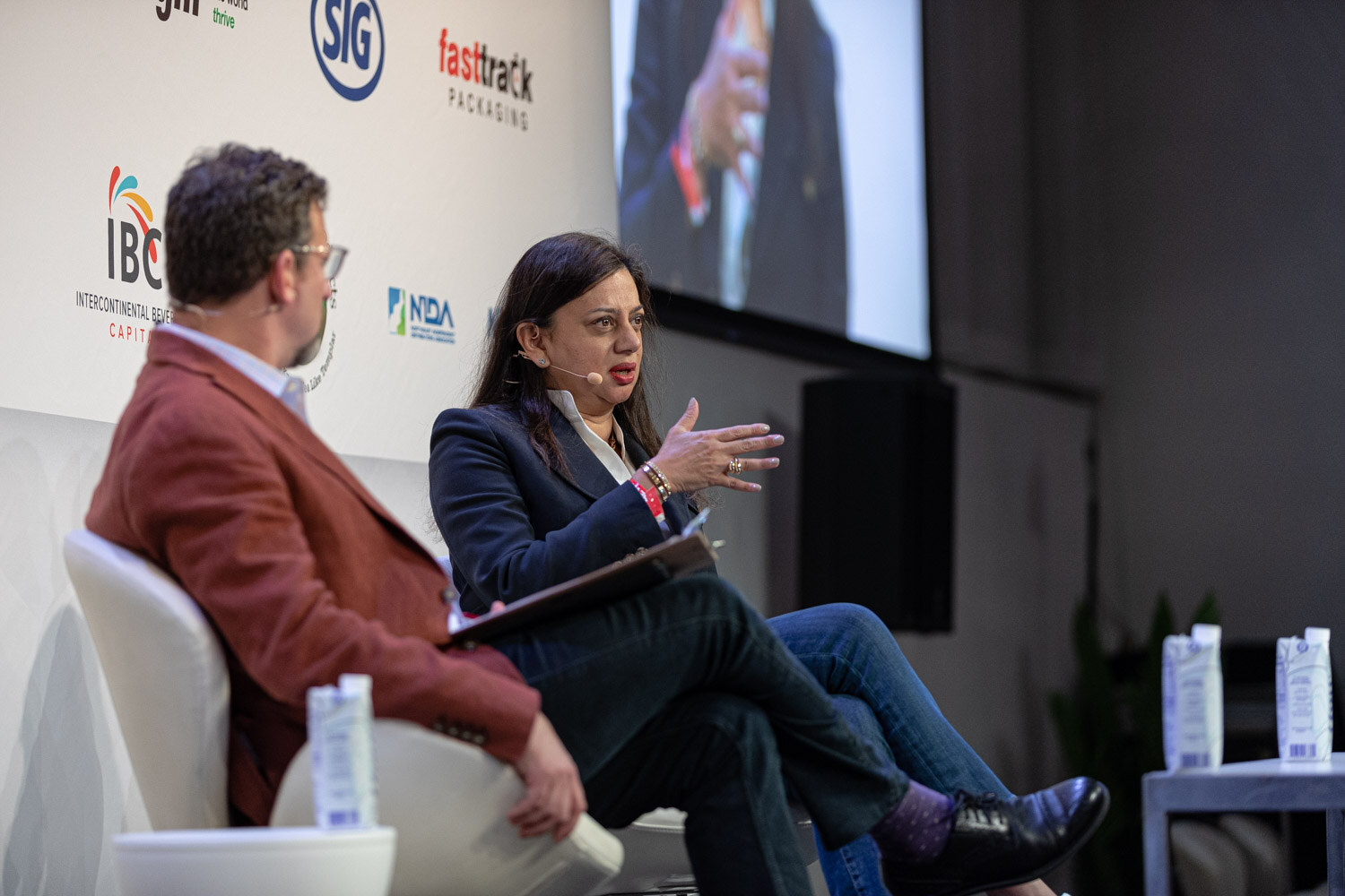 A man in a rust jacket and a woman in a navy blazer talk on stage while a screen behind them shows another angle.
