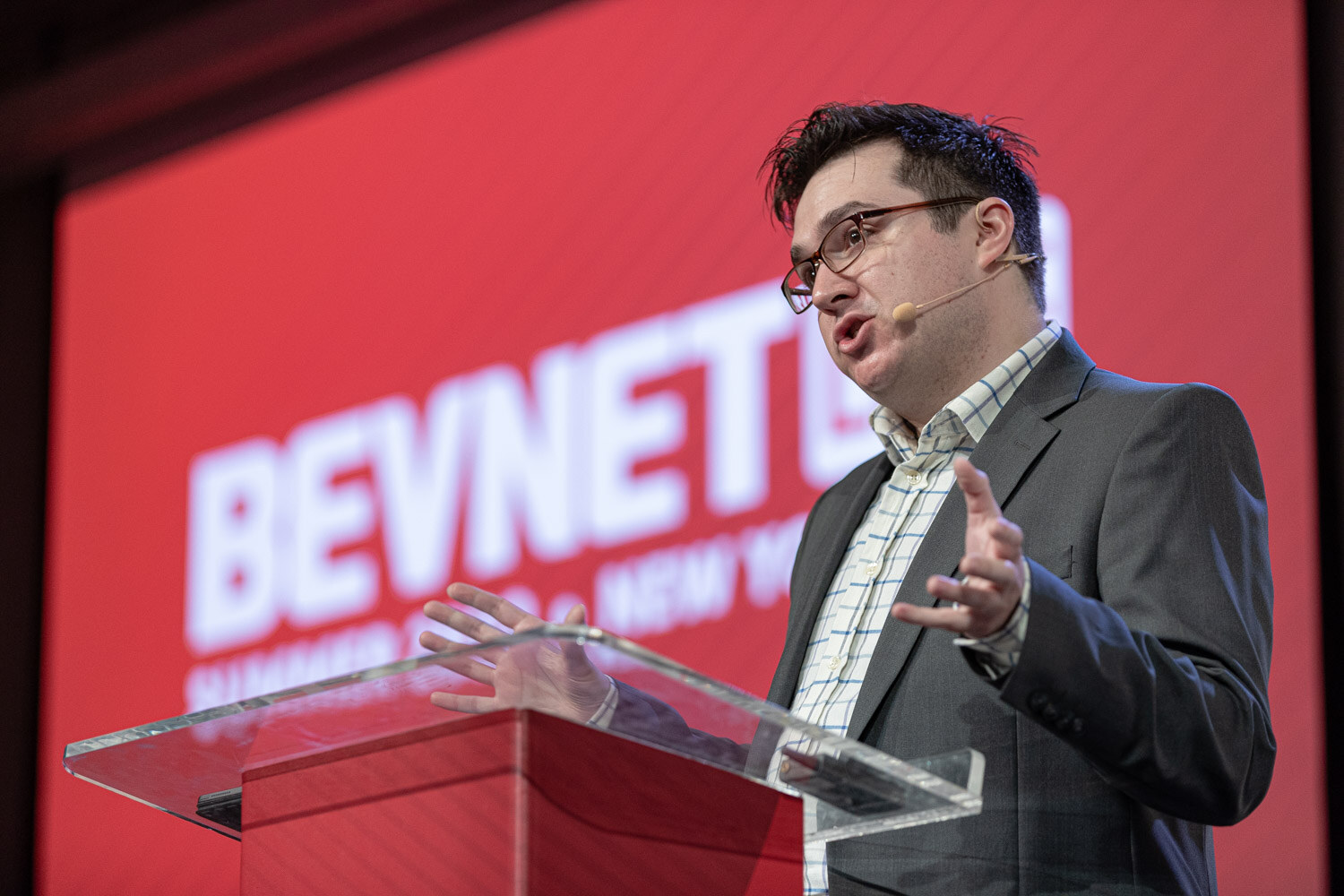 A man in glasses and a gray jacket gestures behind a clear podium in front of a red BevNET Live screen.