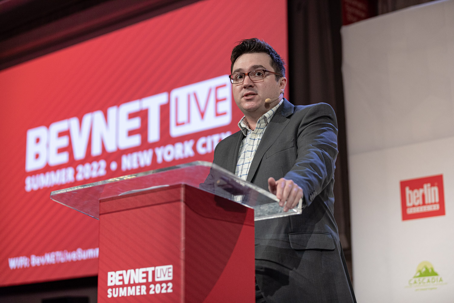 A man in glasses and a gray jacket speaks behind a clear podium with a red screen reading BevNET Live Summer 2022 New York City.