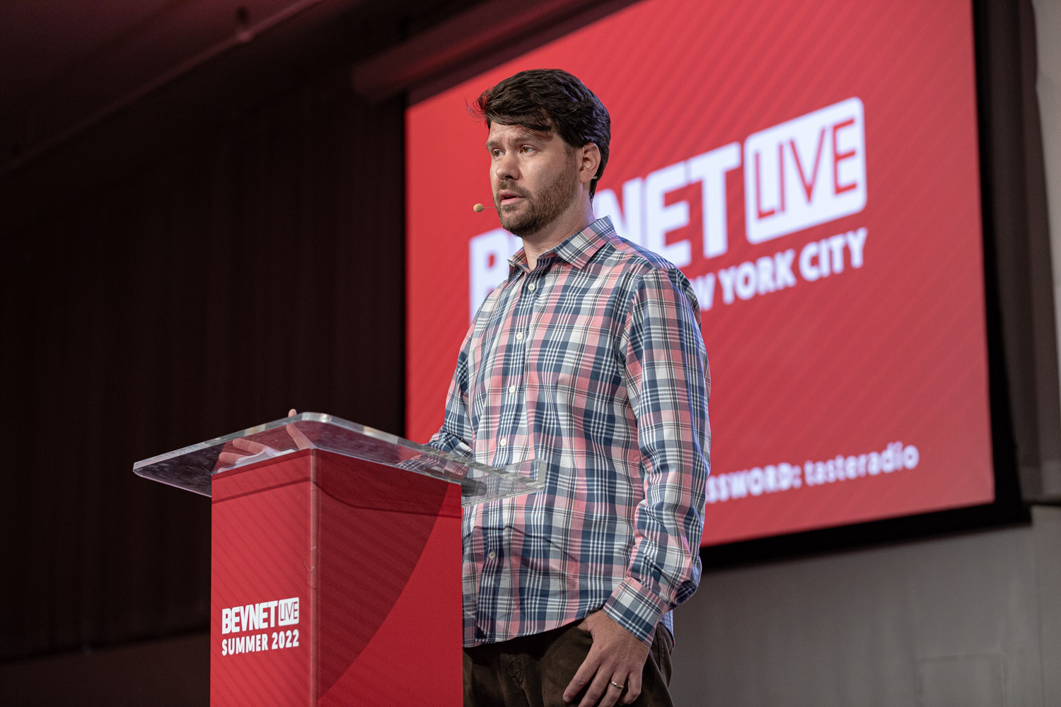 A man in a plaid shirt speaks into a headset microphone at a clear podium in front of a red BevNET Live screen.
