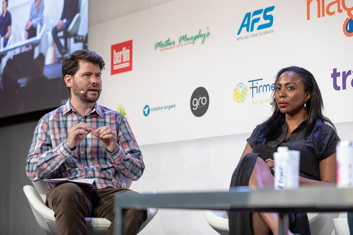 A man in a plaid shirt with a notepad gestures at a woman in a black dress, who sits beside him in a white chair.