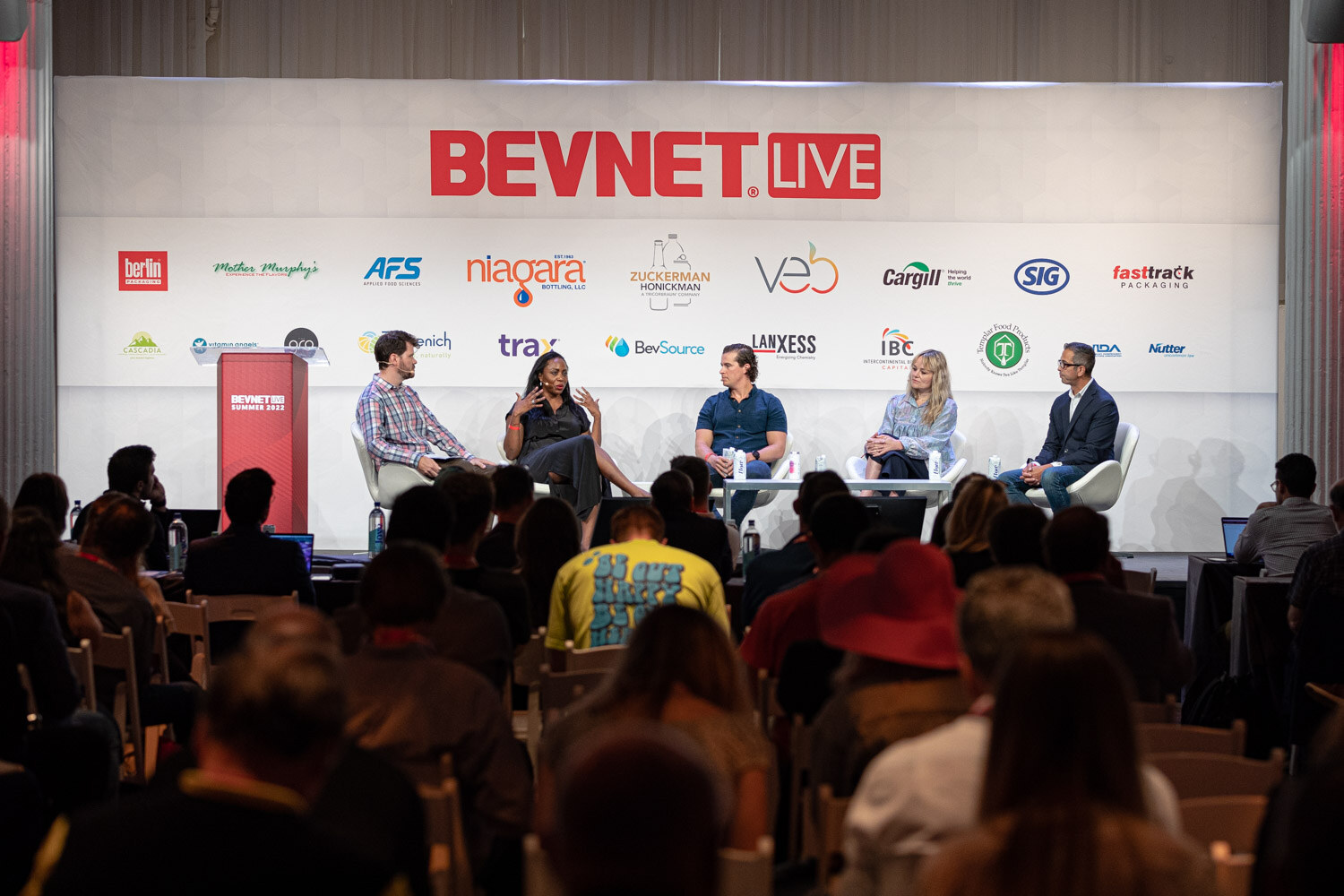 Wide view of five people in white chairs under a BevNET Live backdrop, audience heads in the foreground.