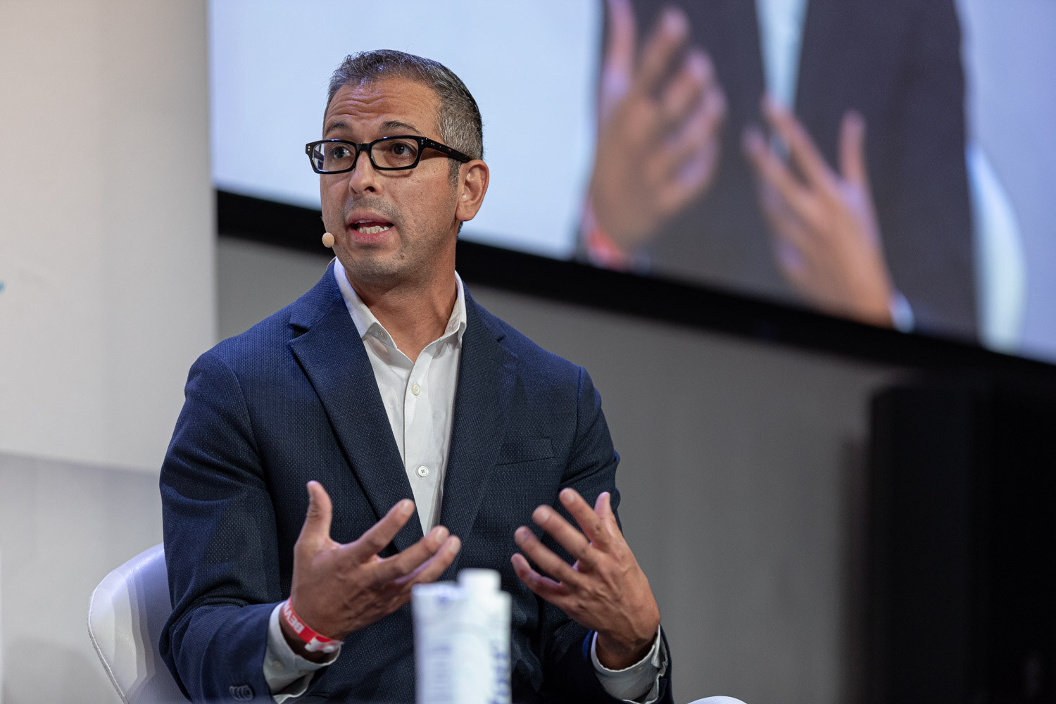 A man with glasses and a navy blazer gestures with both hands, a blurred screen behind him.