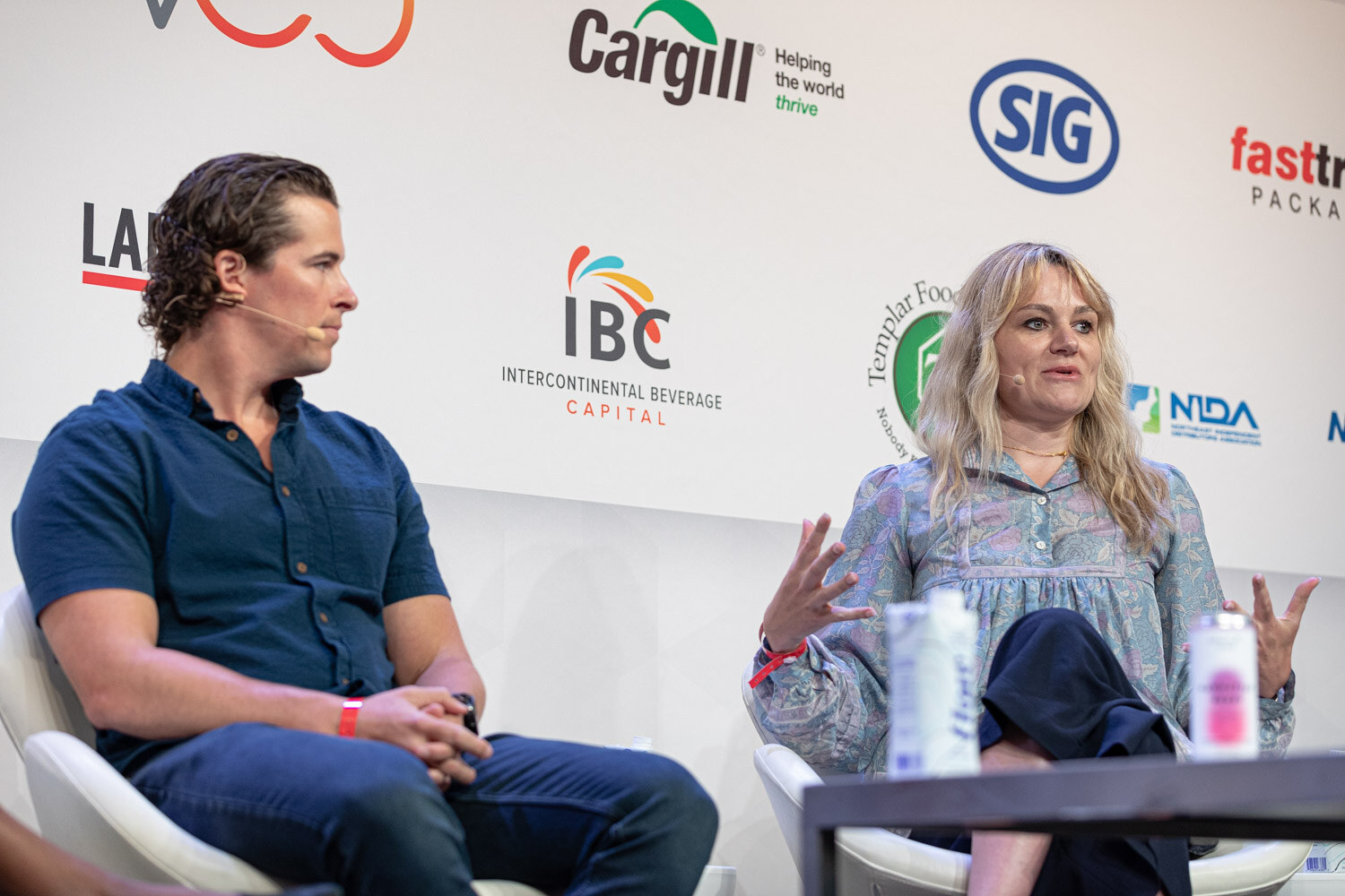 A man in a navy shirt listens while a woman in a floral blouse gestures with both hands, in white chairs in front of sponsor logos.