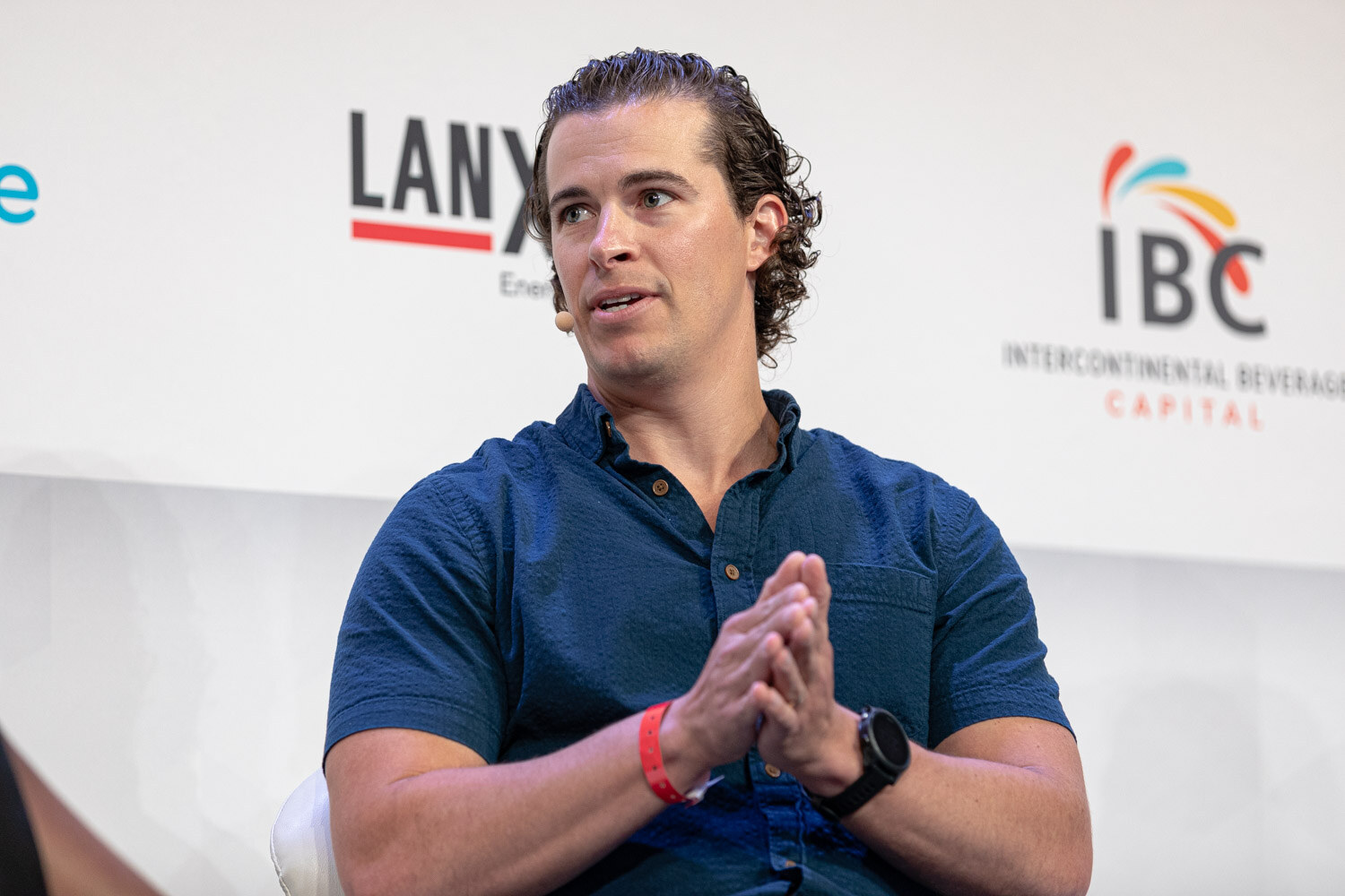 A man with dark curly hair in a navy shirt and red wristband looks to his side, with IBC and Lanxess signs behind him.
