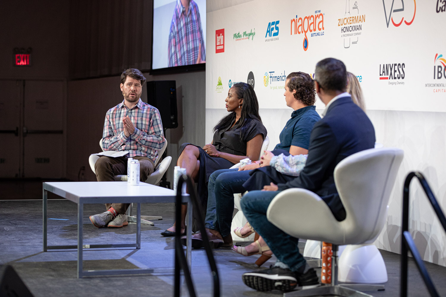 Five people seated on stage in white chairs with a low table in front, a screen at upper left showing the speaker.
