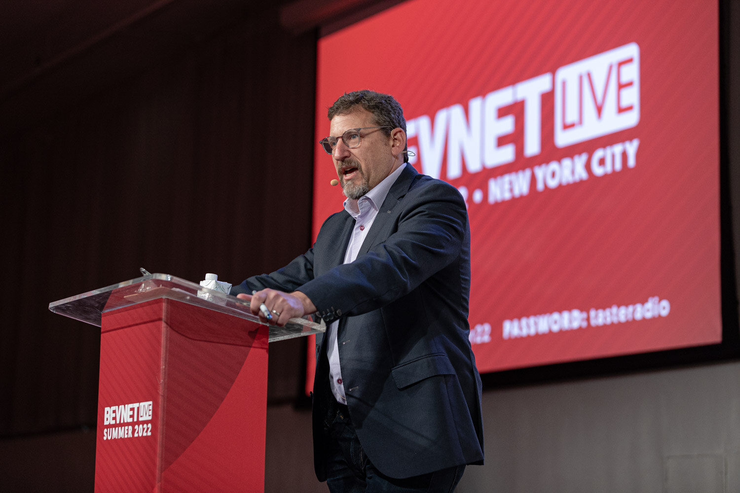 A man with glasses and a gray beard in a navy blazer speaks at a clear podium in front of a red BevNET Live screen.