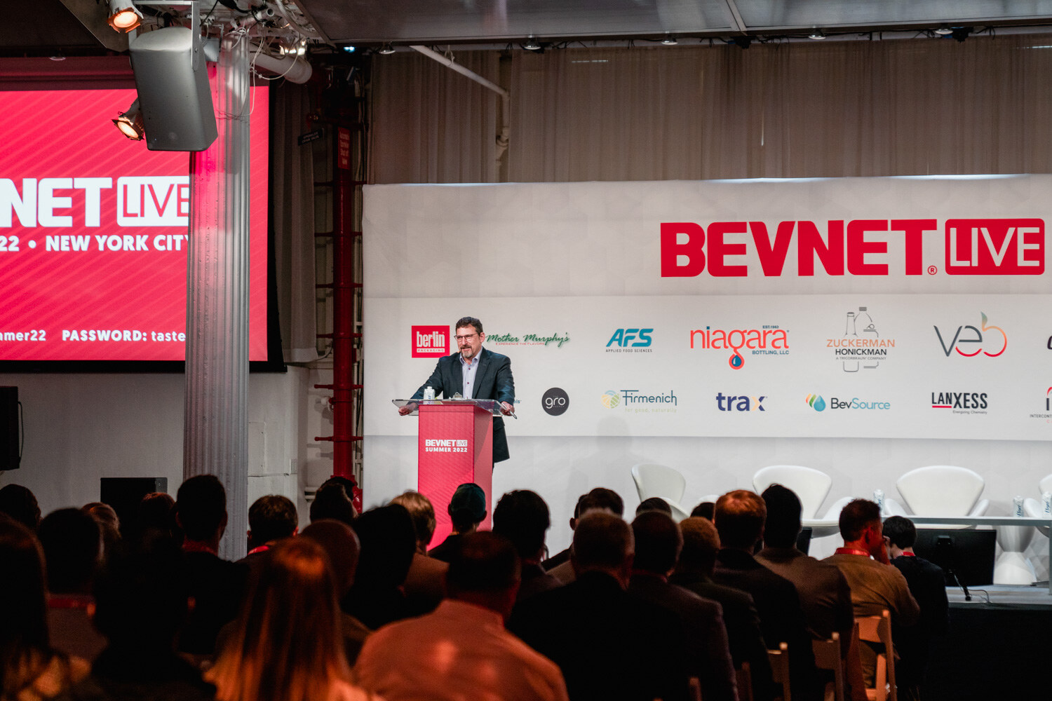 Wide view of a man at a red podium on the BevNET Live stage with silhouetted audience members in front.
