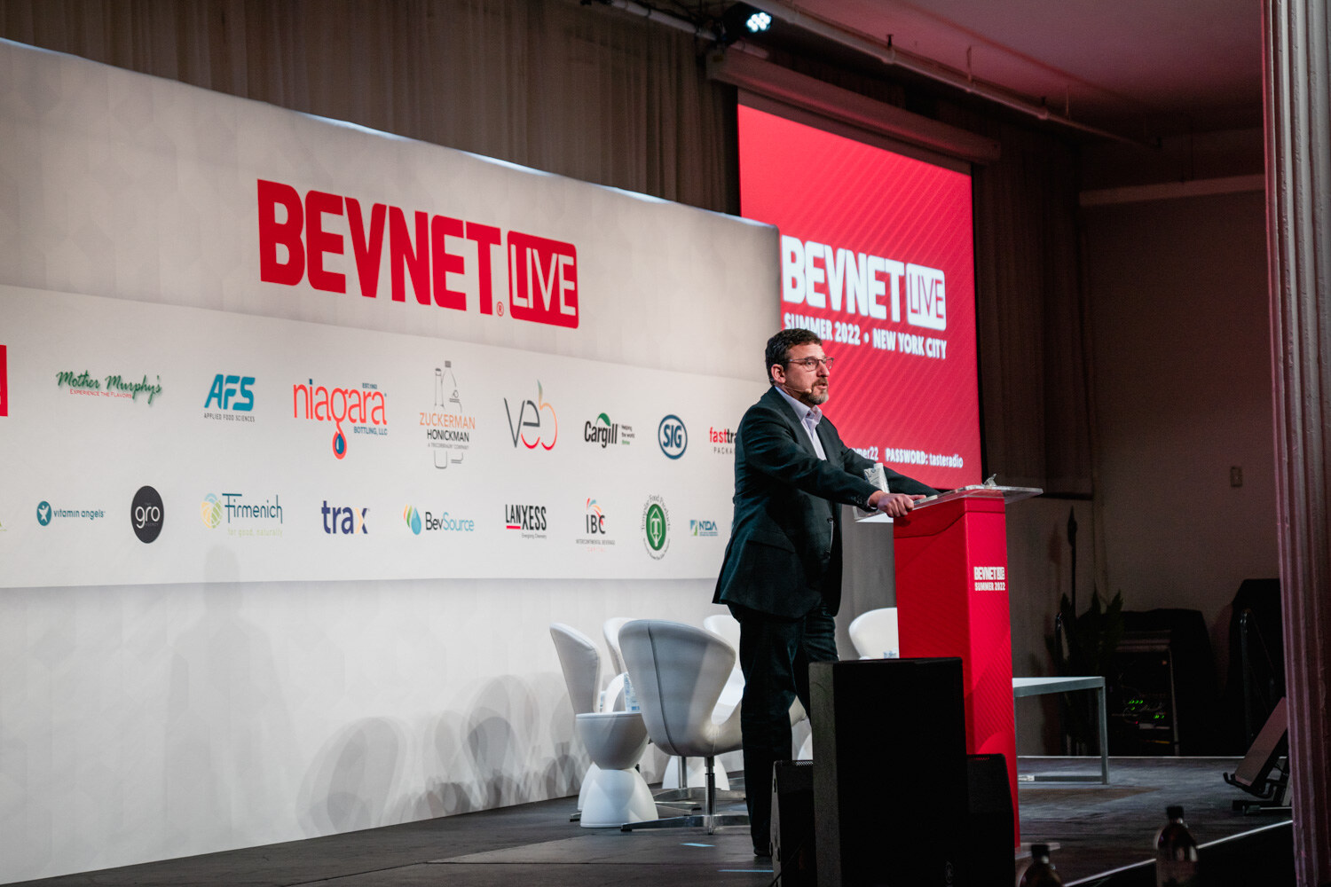 A man in a navy blazer stands at a red podium on a stage with white chairs and a red BevNET Live screen at right.