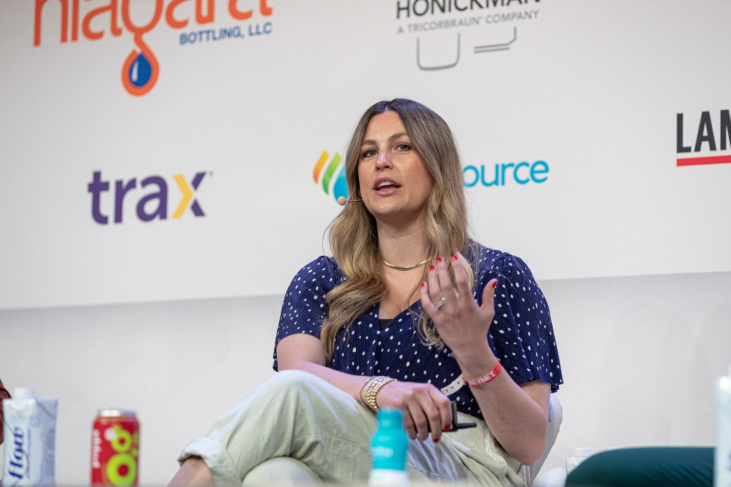 A woman in a polka-dot blouse and cream pants speaks with her left hand raised, with Trax and BevSource logos behind her.