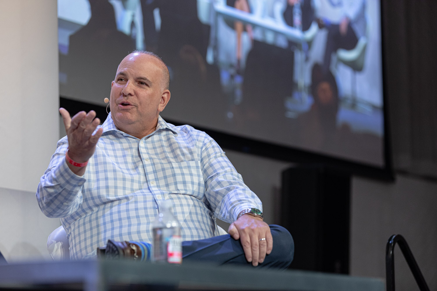 A man in a light blue checked shirt sits with his legs crossed, gesturing with his right hand, a big screen behind him.